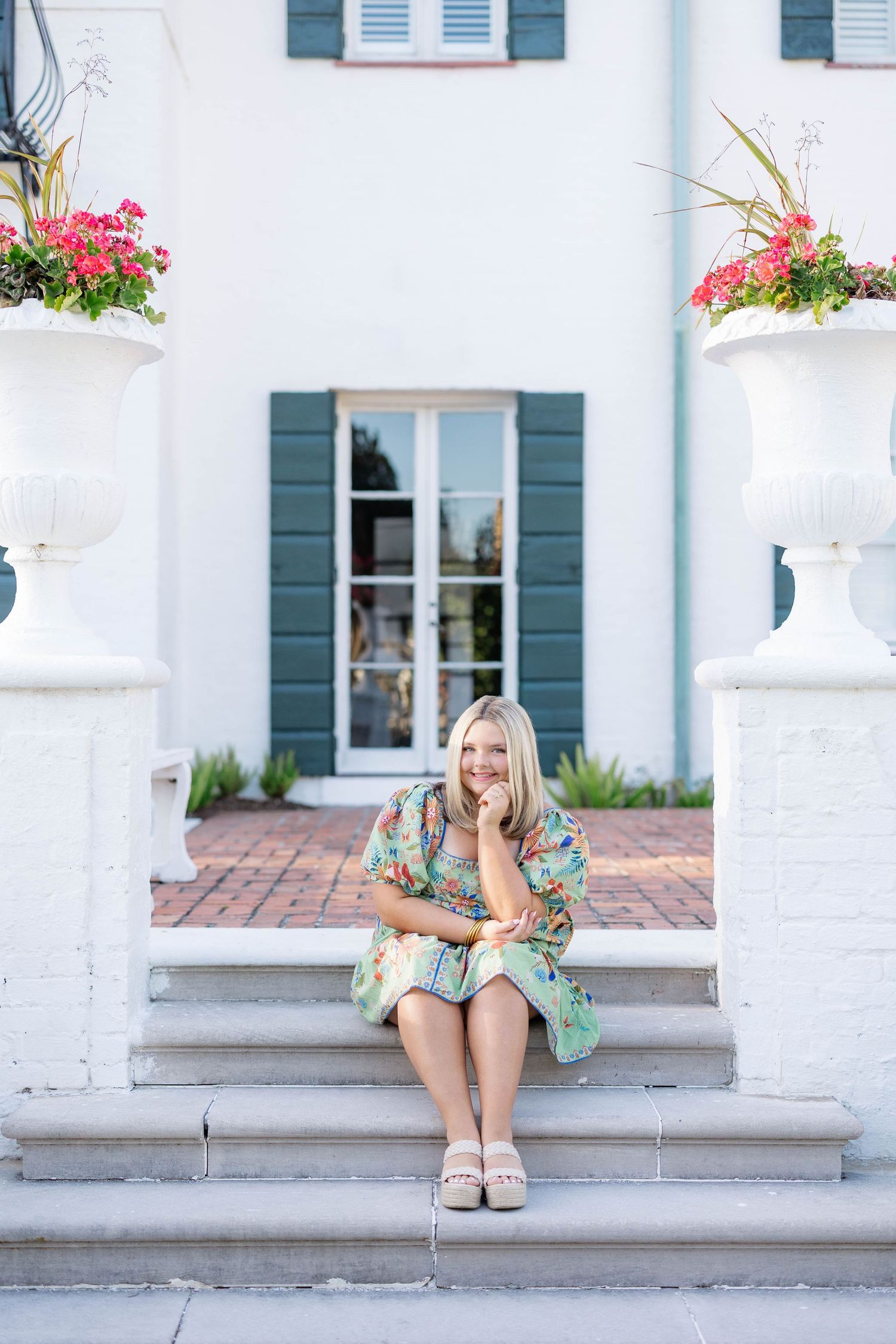 Smiling blonde woman in floral dress sitting on white steps with pink flower urns outside a white building.