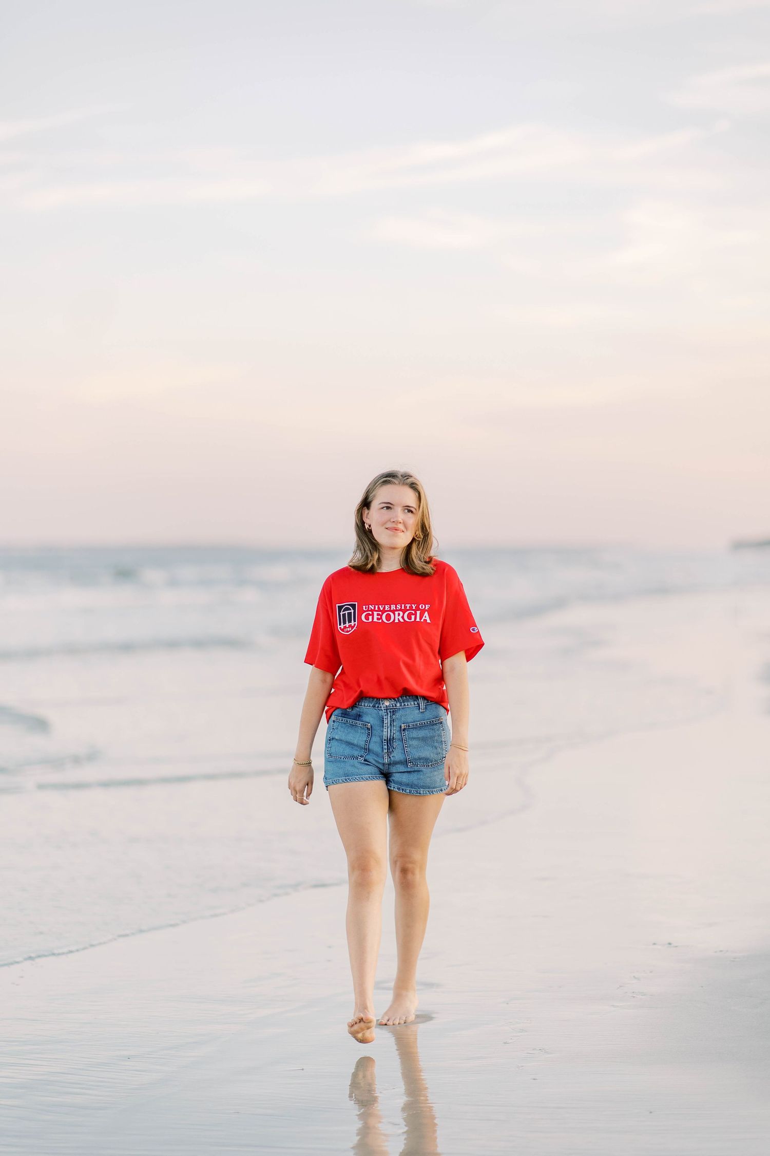 Young woman in red University of Georgia t-shirt and denim shorts walking barefoot on a beach at sunset.