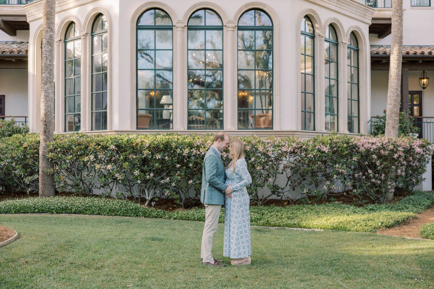 Couple embracing on lush green lawn in front of elegant building with arched windows during golden hour.