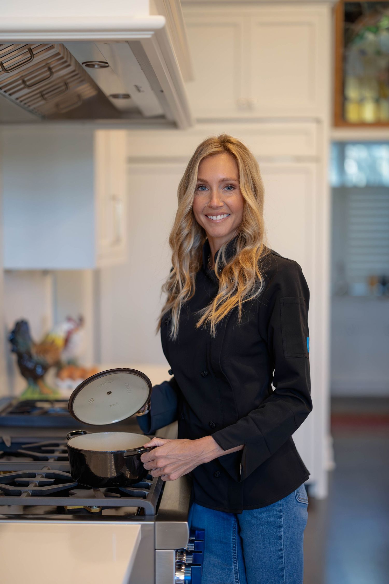 Smiling blonde woman in black chef jacket lifting lid off pot on stainless steel stove in bright white kitchen.