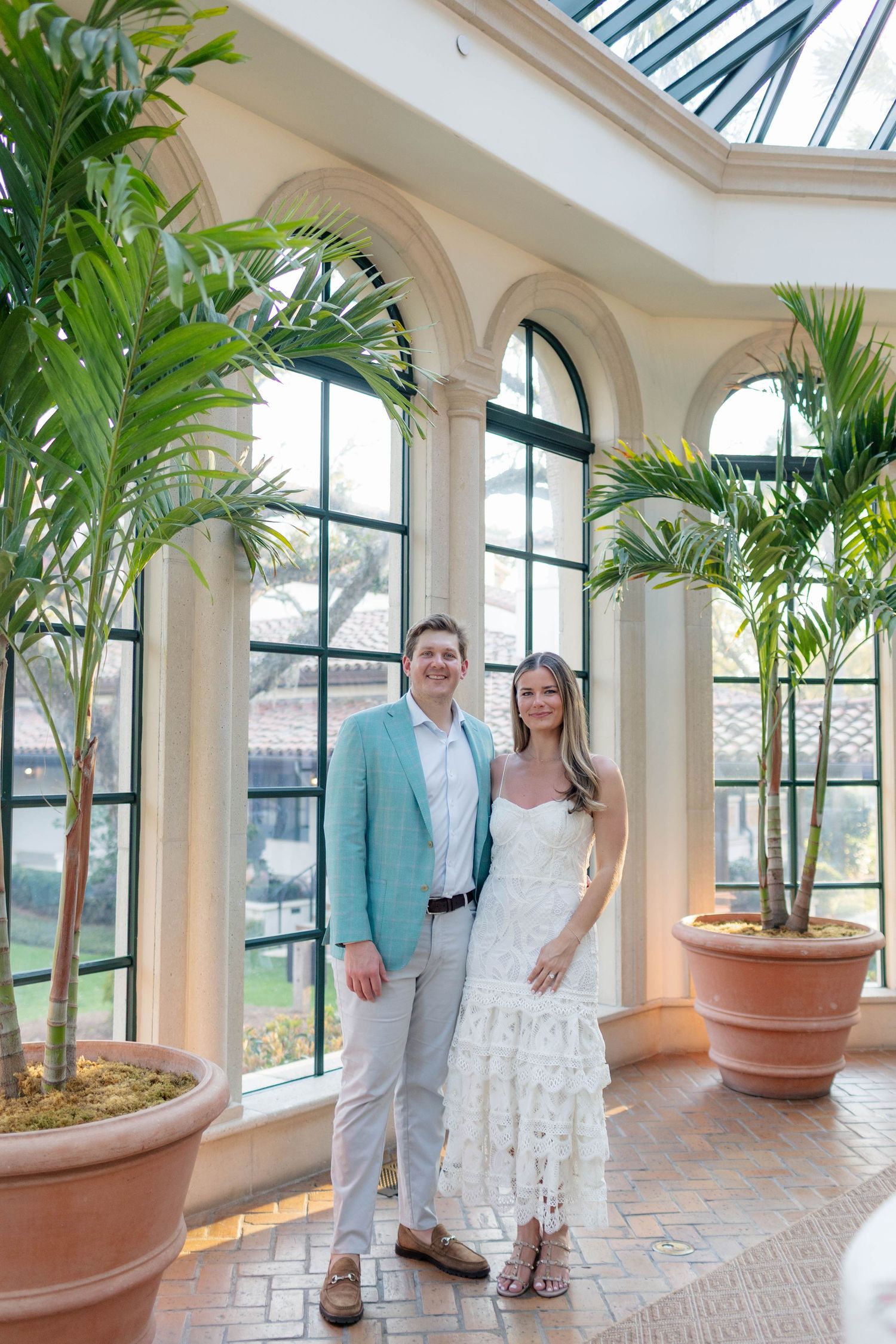 Couple posing in elegant conservatory with arched windows, palm trees, and skylight, man in blue blazer, woman in white lace dress.