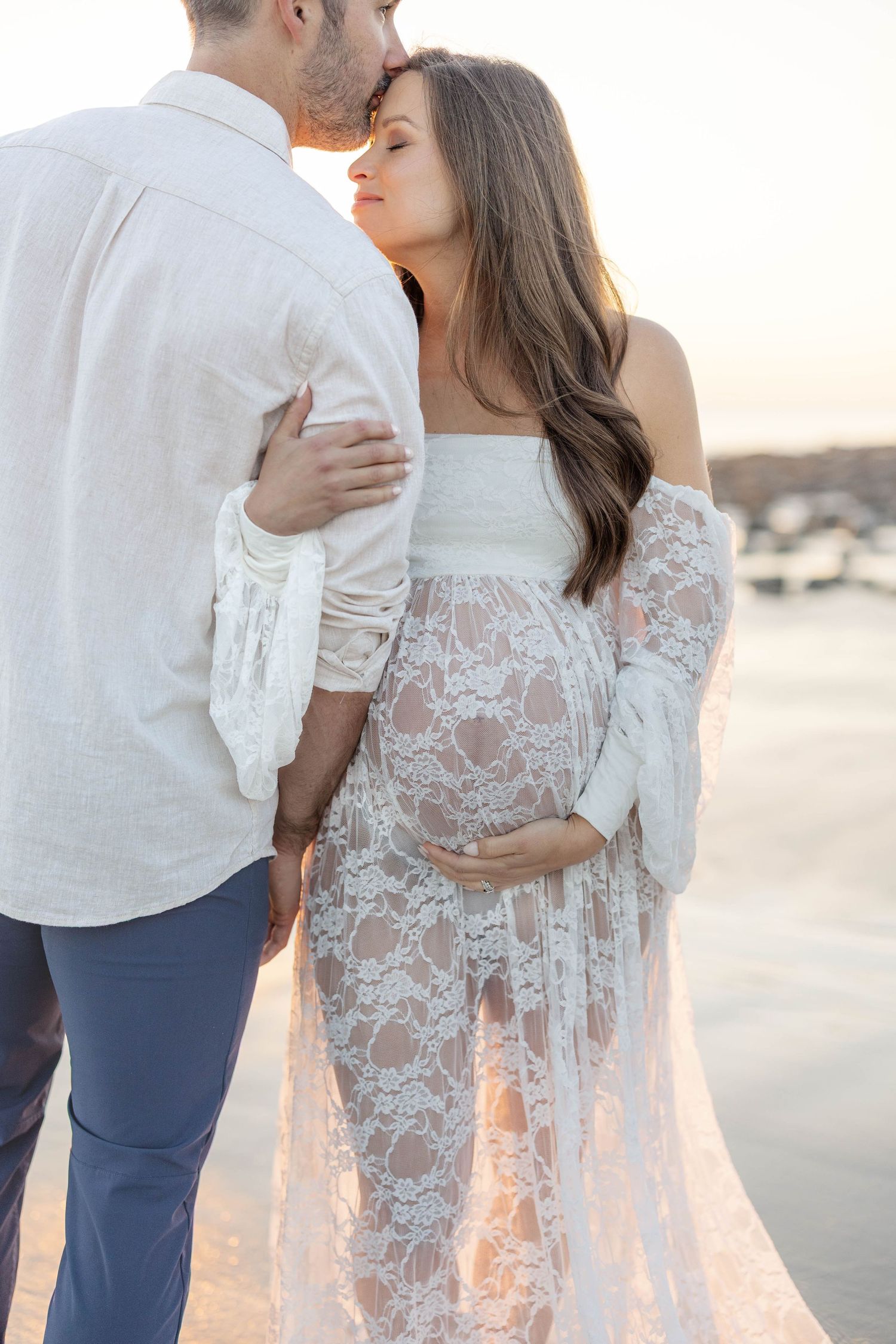 Pregnant woman in white lace maternity gown embraces partner on beach at golden sunset.