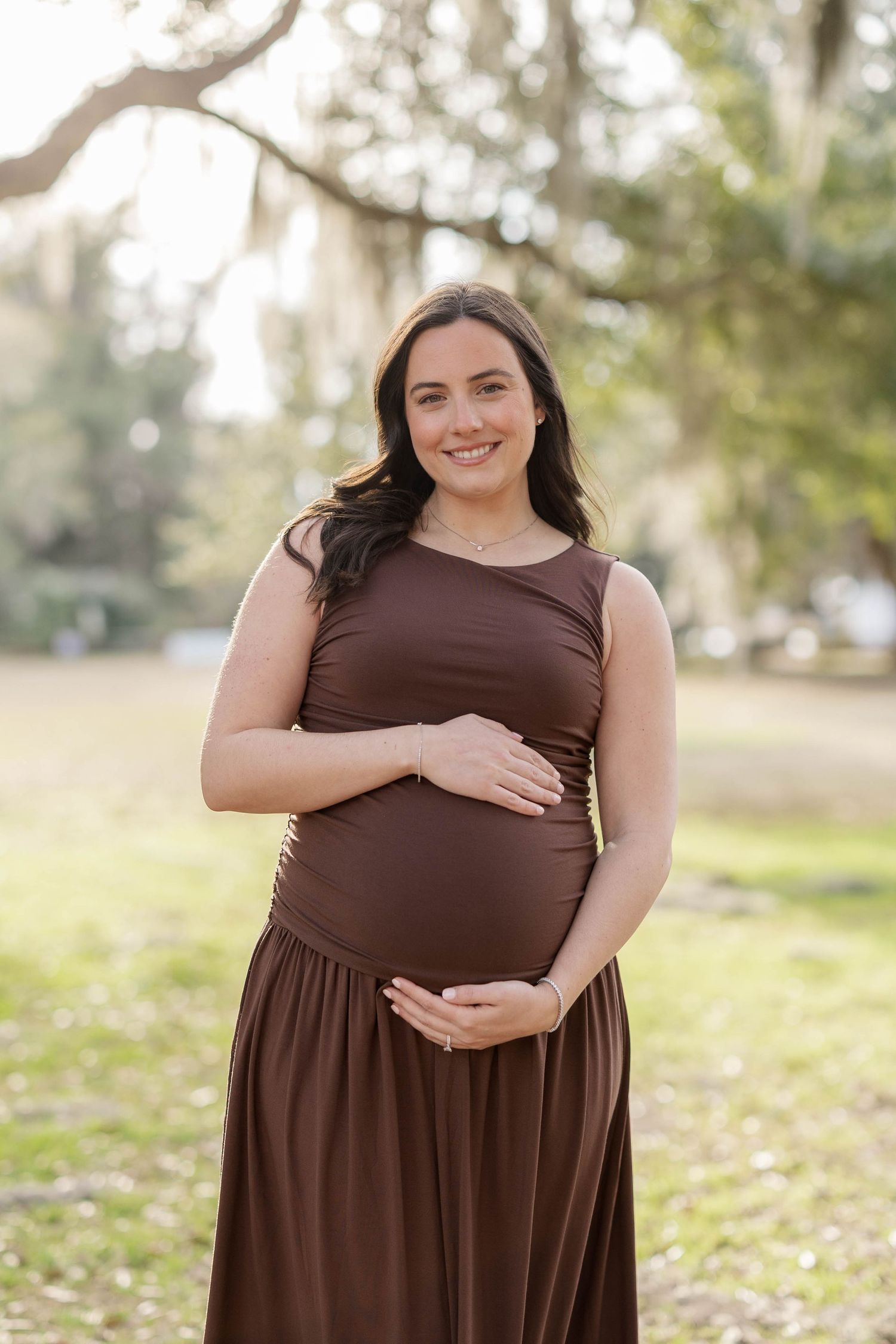 Smiling pregnant woman in brown maxi dress cradling baby bump outdoors in a sunlit park setting.