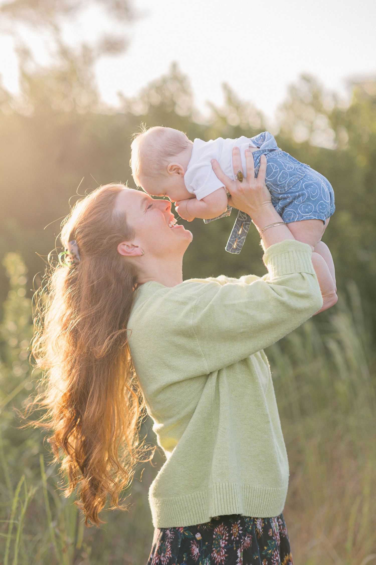 A joyful mother lifts her baby in the air outdoors in golden sunlight, both smiling in a lush green field.