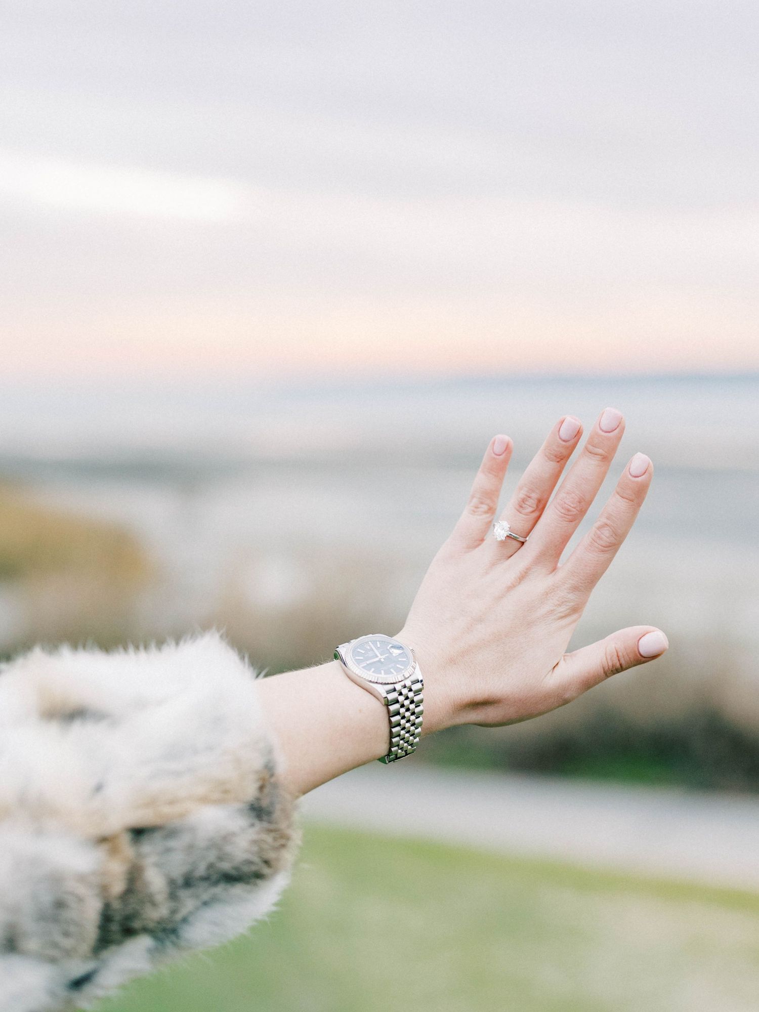 A woman's hand wearing a gold ring and silver watch with a fur coat sleeve, raised against a serene ocean sunset backdrop.