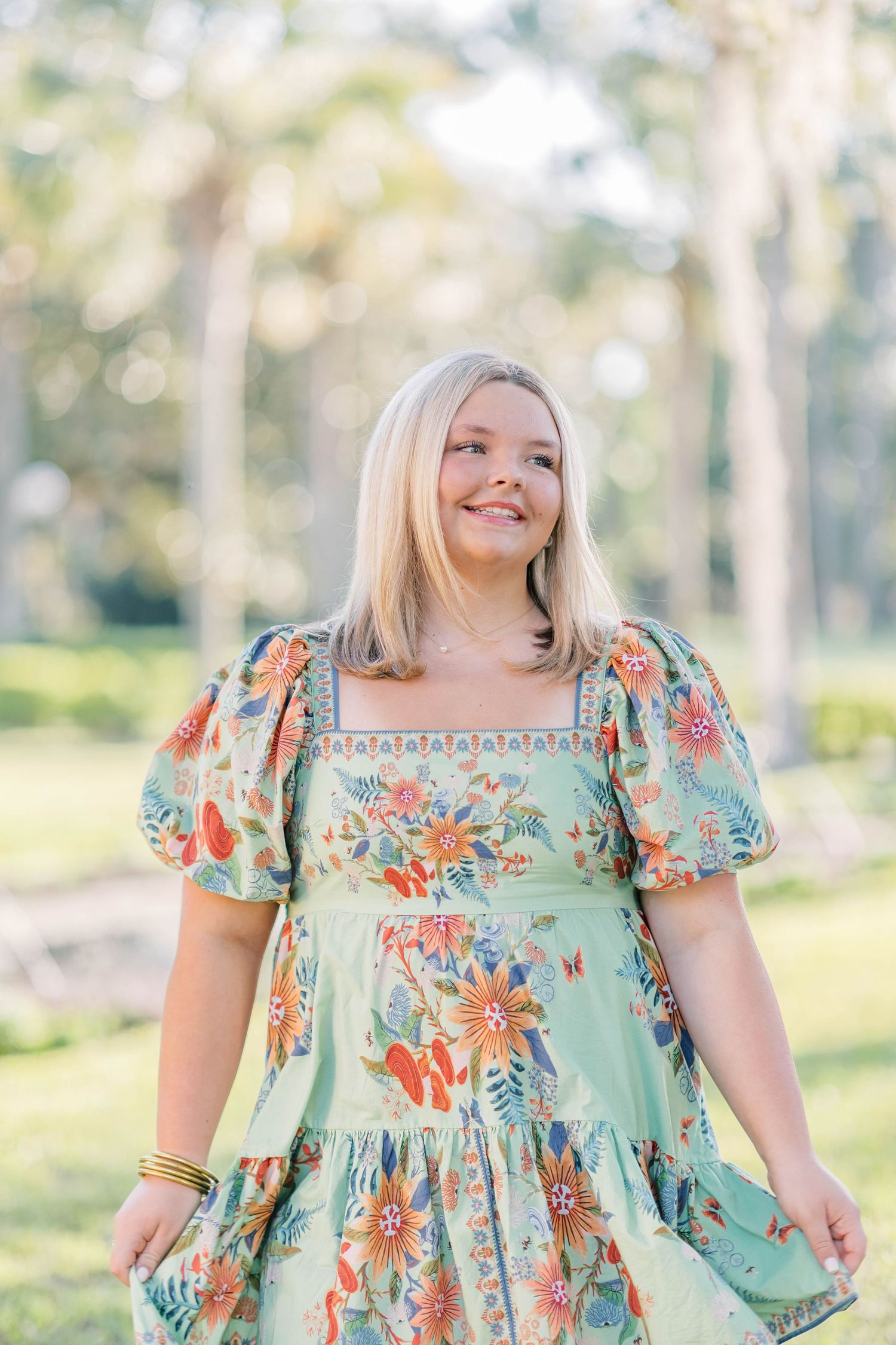 Smiling blonde woman in a floral mint green dress standing outdoors among tall trees in a park.
