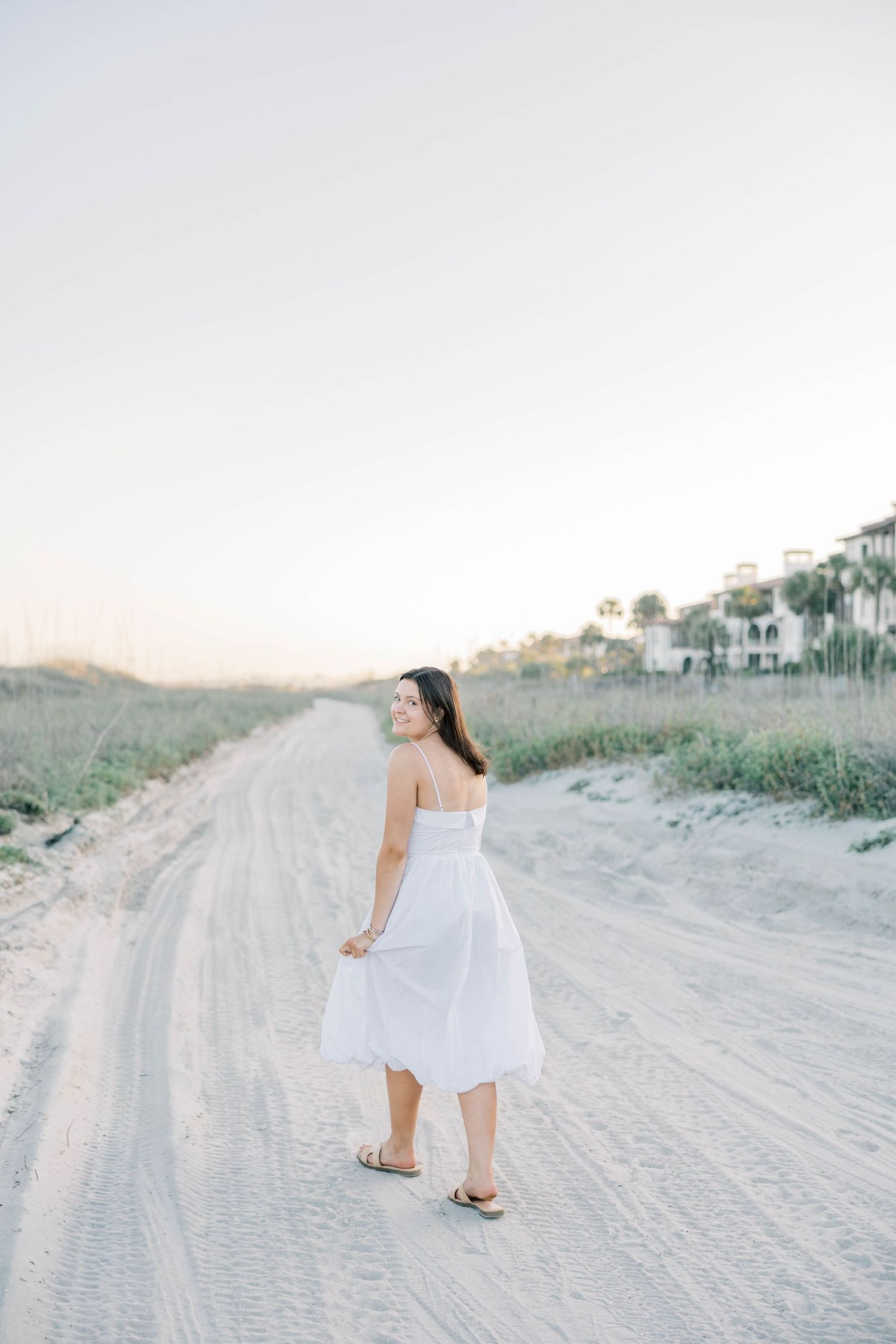 Woman in white sundress walking on sandy beach path at golden hour, glancing back over her shoulder.