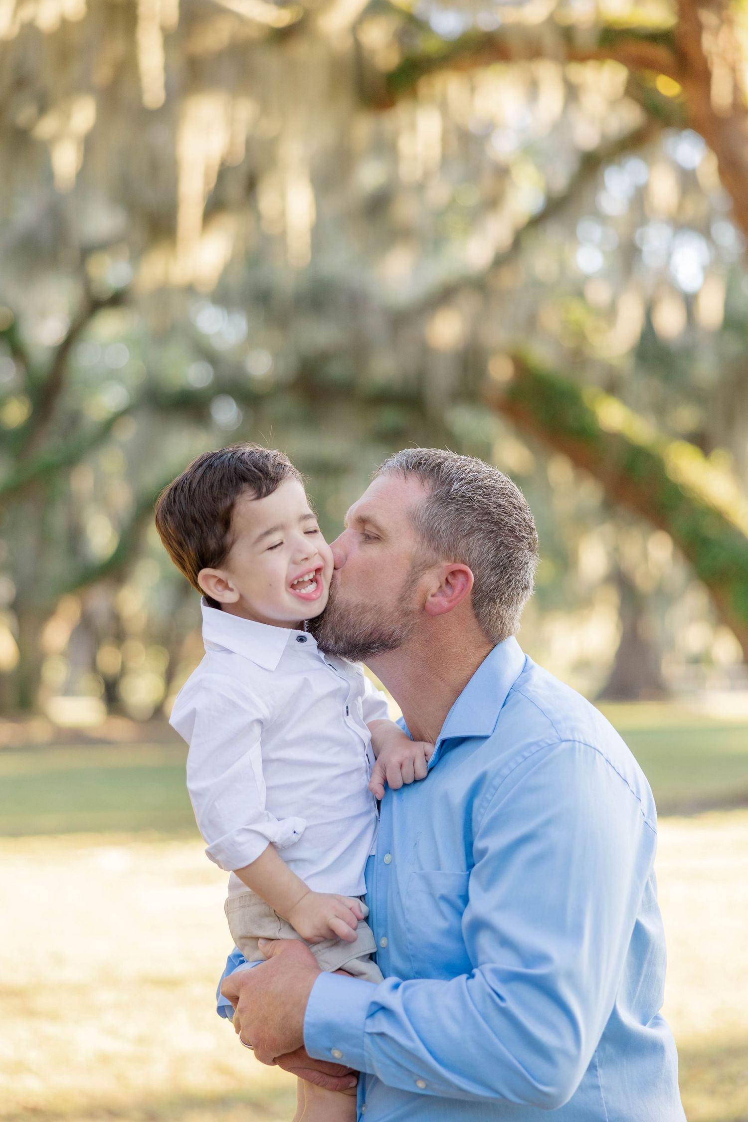 Loving father in blue shirt kissing smiling young son while holding him outdoors.