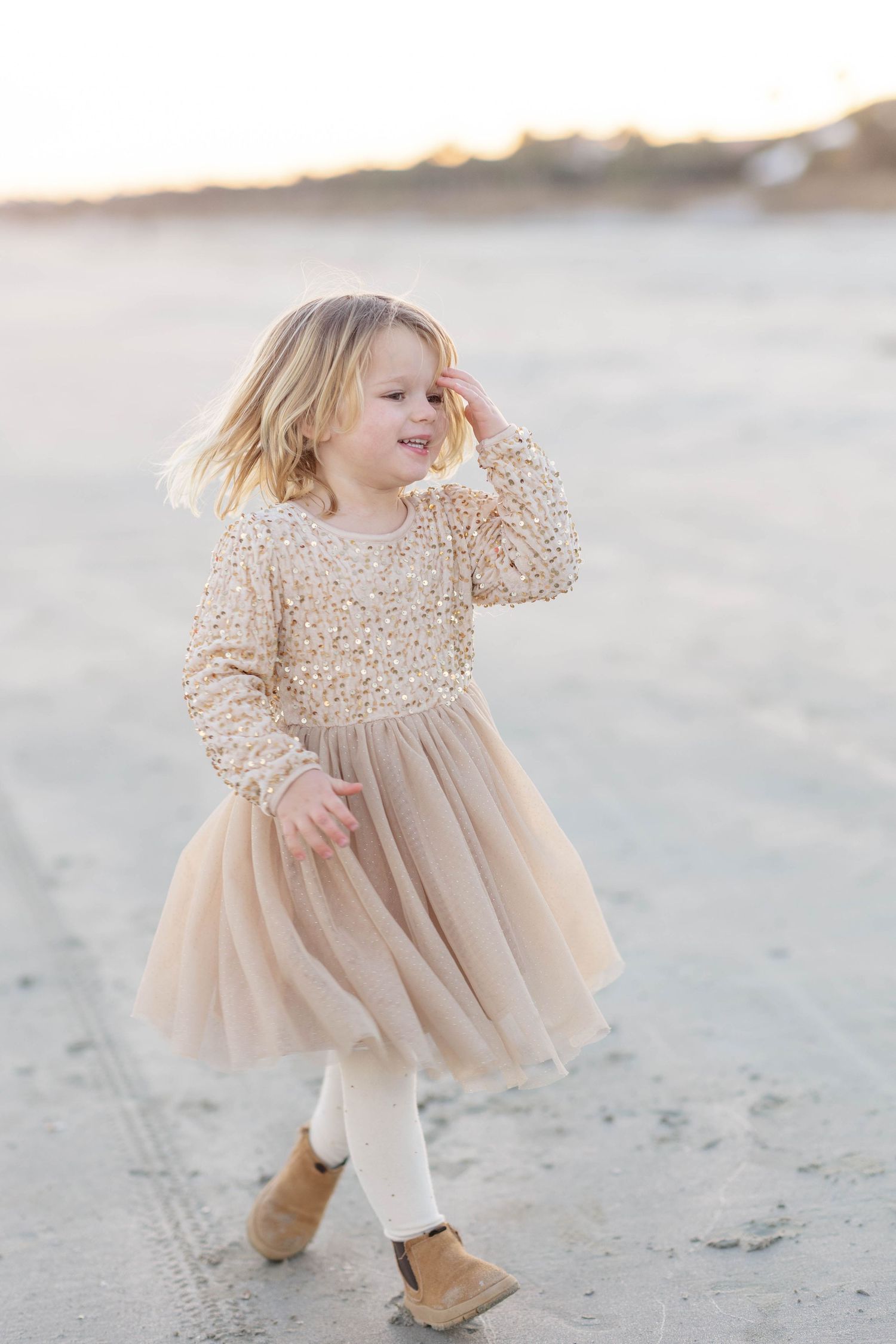 Young girl in a sparkly gold tulle dress walking on a sandy beach at sunset.
