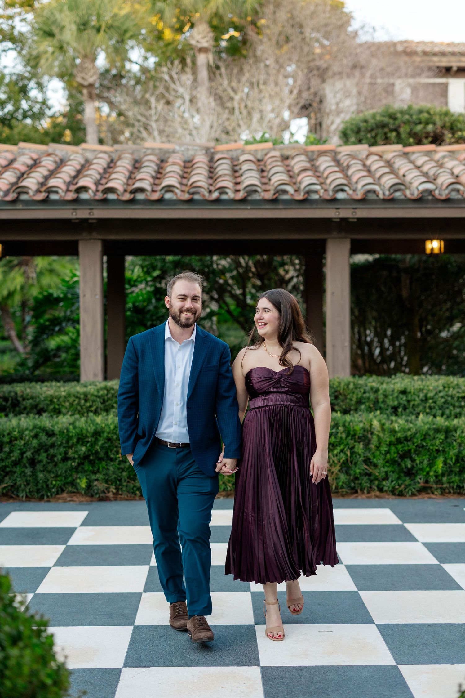 Couple walking hand-in-hand on checkered floor, man in blue suit, woman in burgundy dress, Spanish-style building backdrop.