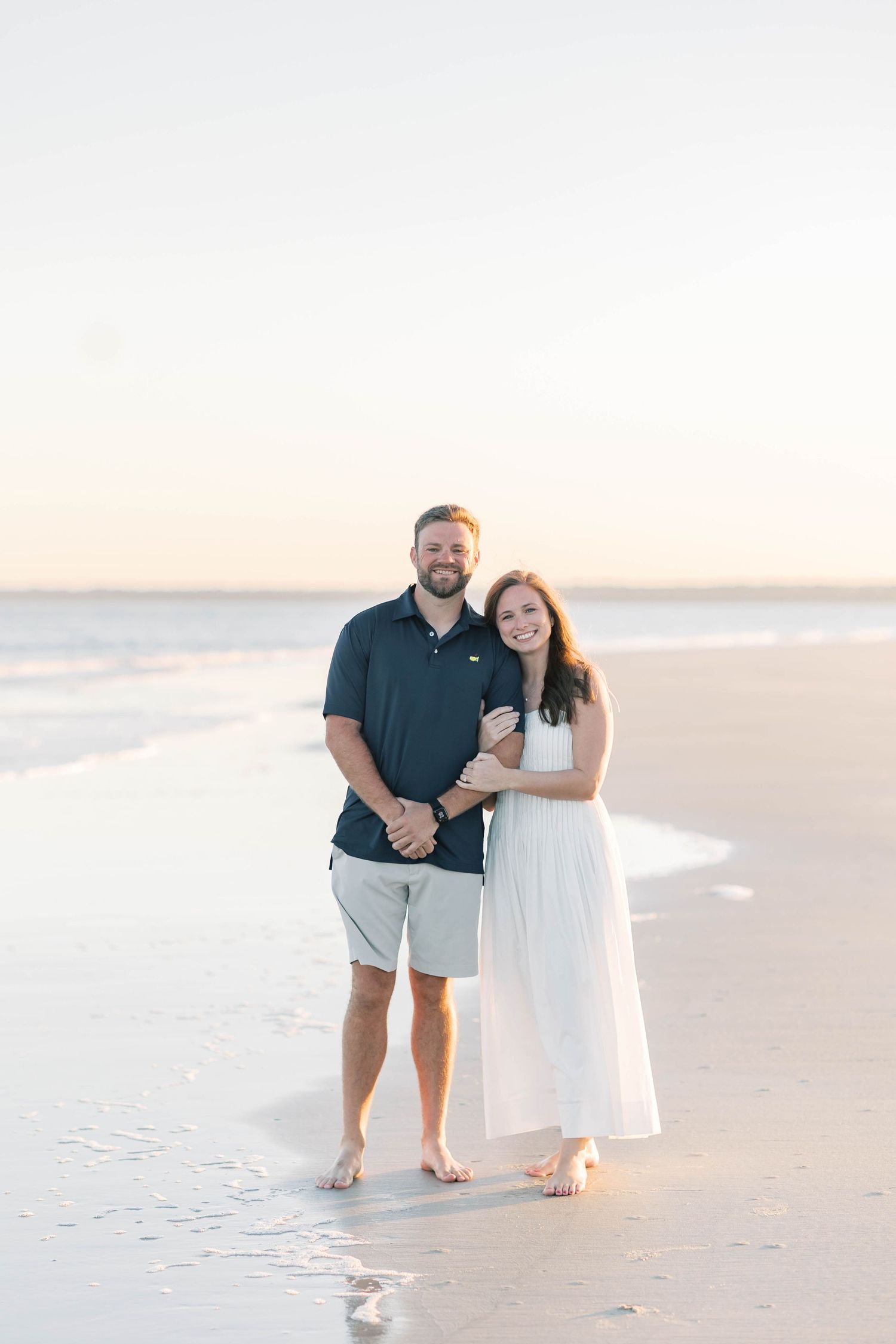 Happy couple embracing on a serene beach at sunset, man in navy polo and khaki shorts, woman in flowing white maxi dress.
