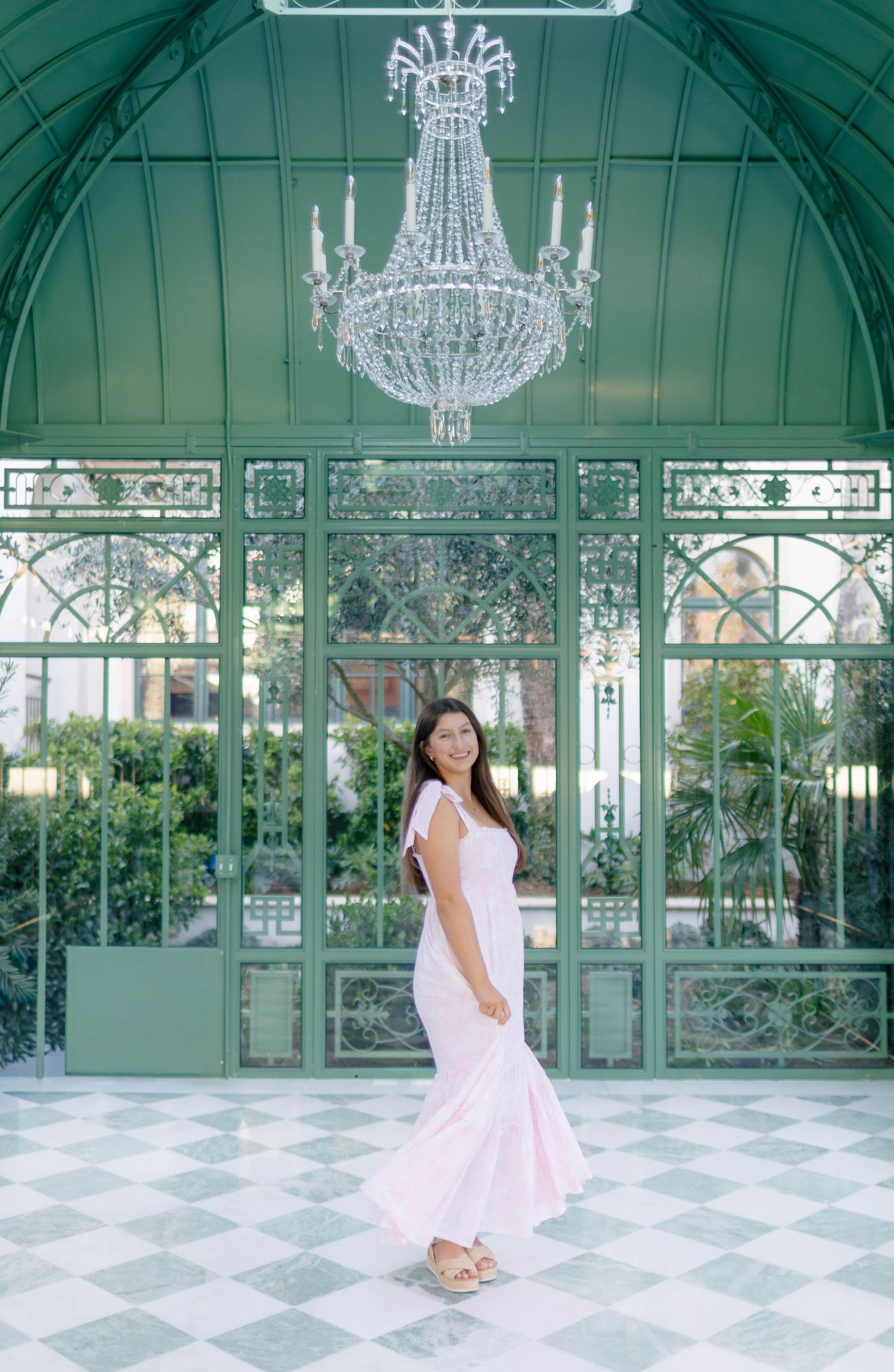 Woman in pink maxi dress posing under crystal chandelier in elegant green conservatory.