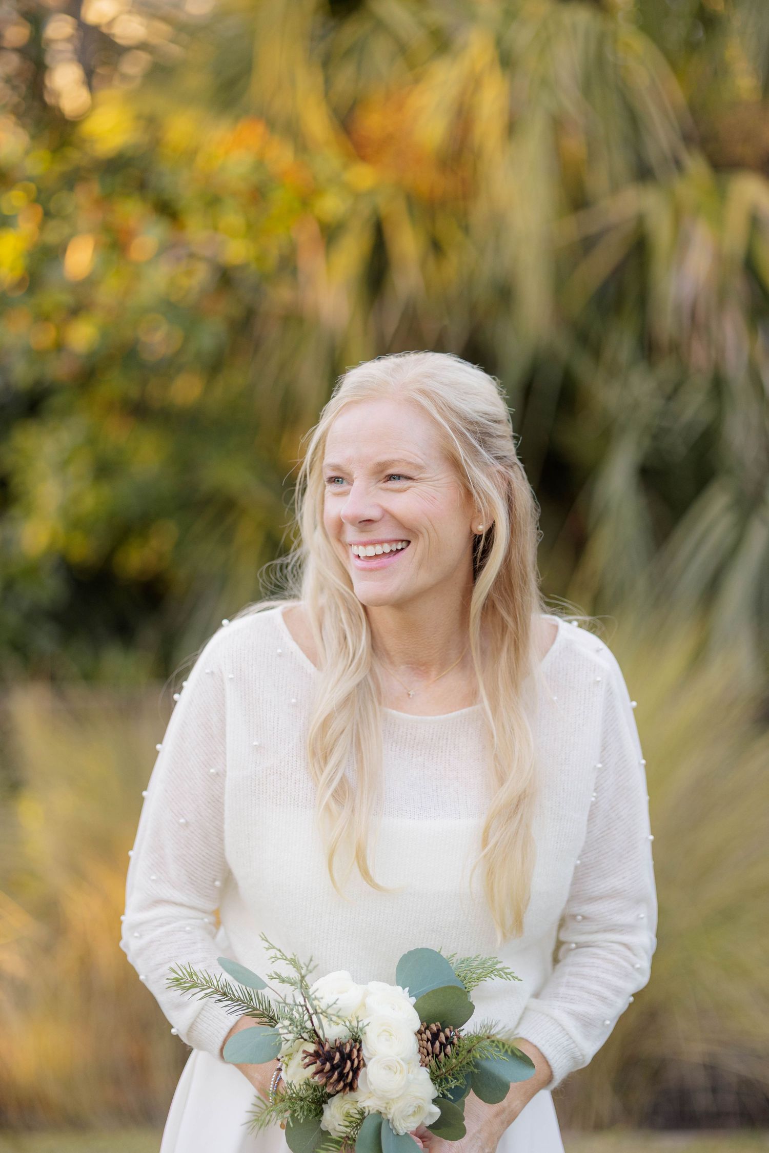 A joyful bride in a white dress holds a winter bouquet with pinecones and eucalyptus against a natural backdrop.