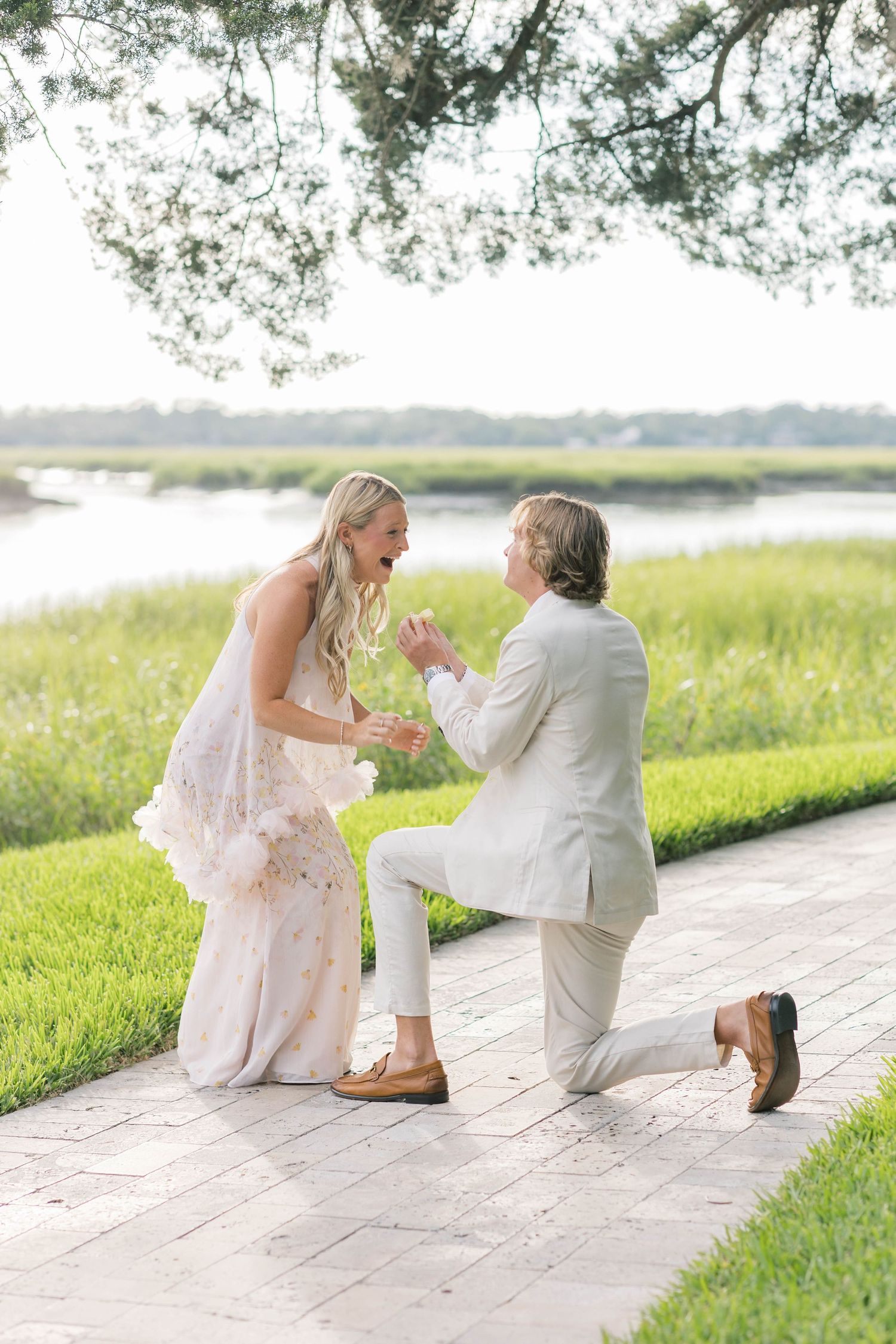 Man in white suit proposes on one knee to surprised blonde woman in floral dress by a scenic waterway.