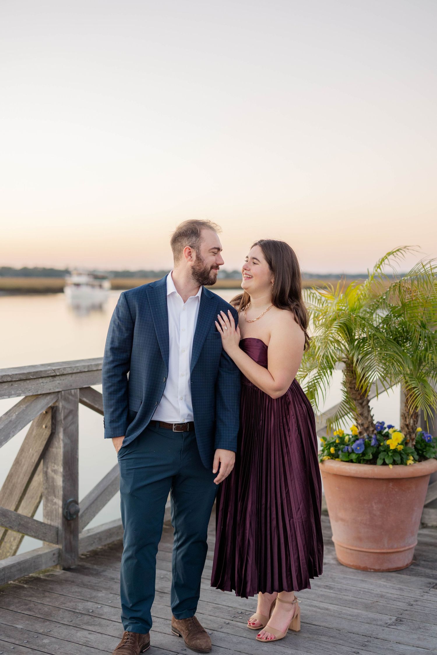 Couple in elegant attire sharing a romantic moment on a waterfront dock at sunset, woman in burgundy dress, man in navy suit.