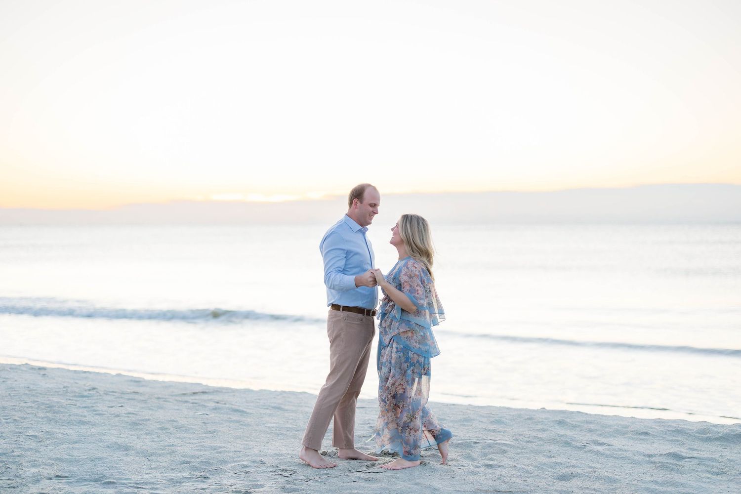 Couple dancing barefoot on a sandy beach at sunset with calm ocean waves in the background.