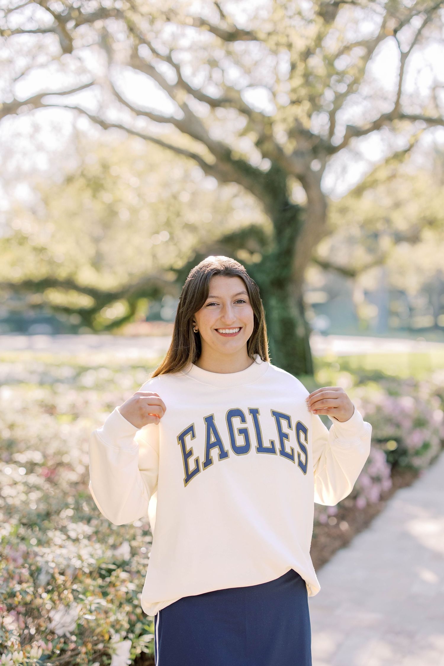 Happy young woman proudly displaying her cream Eagles collegiate sweatshirt while standing in a sunny outdoor garden.