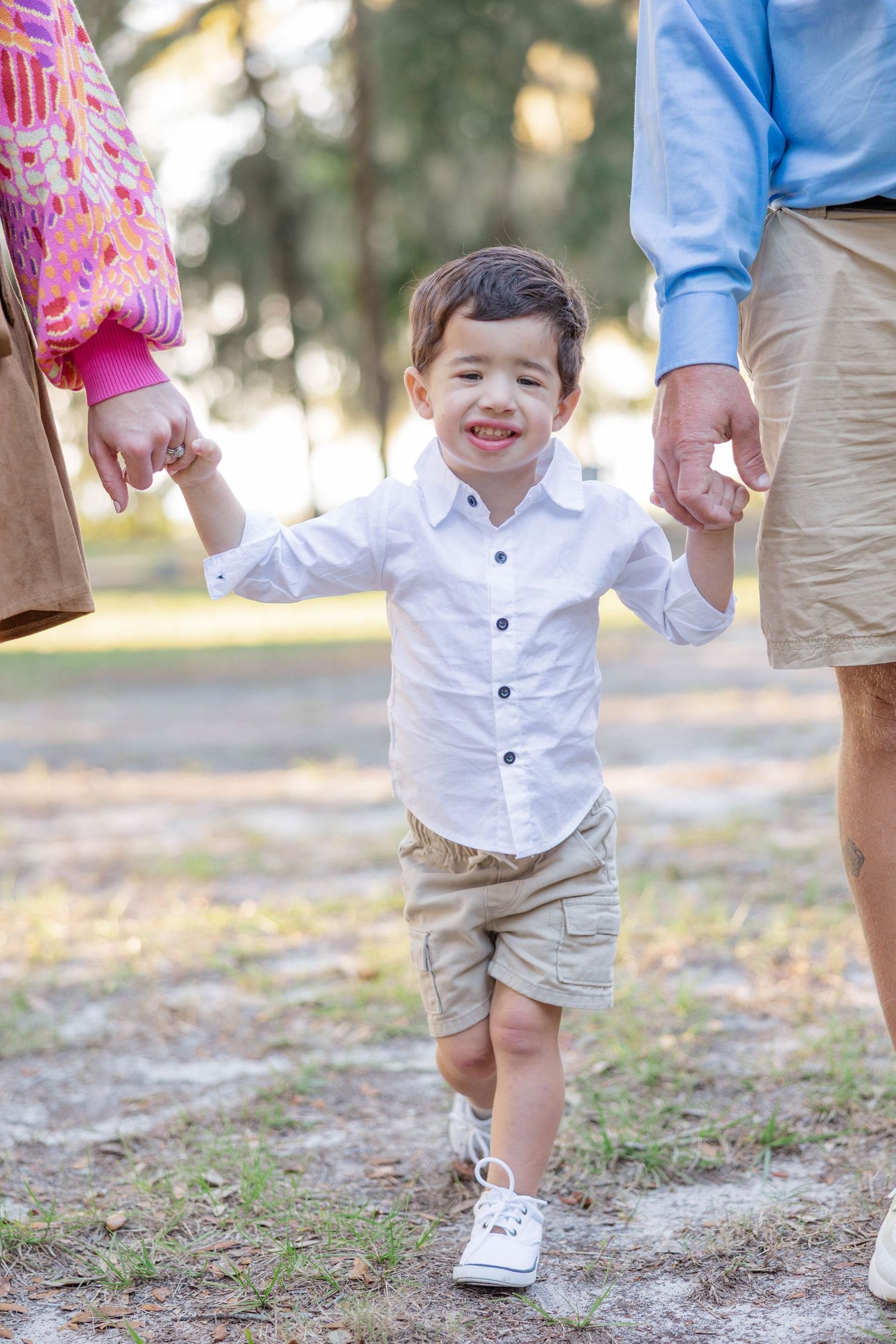 A smiling toddler boy in a white shirt and khaki shorts walks hand-in-hand with parents in a sunny park.