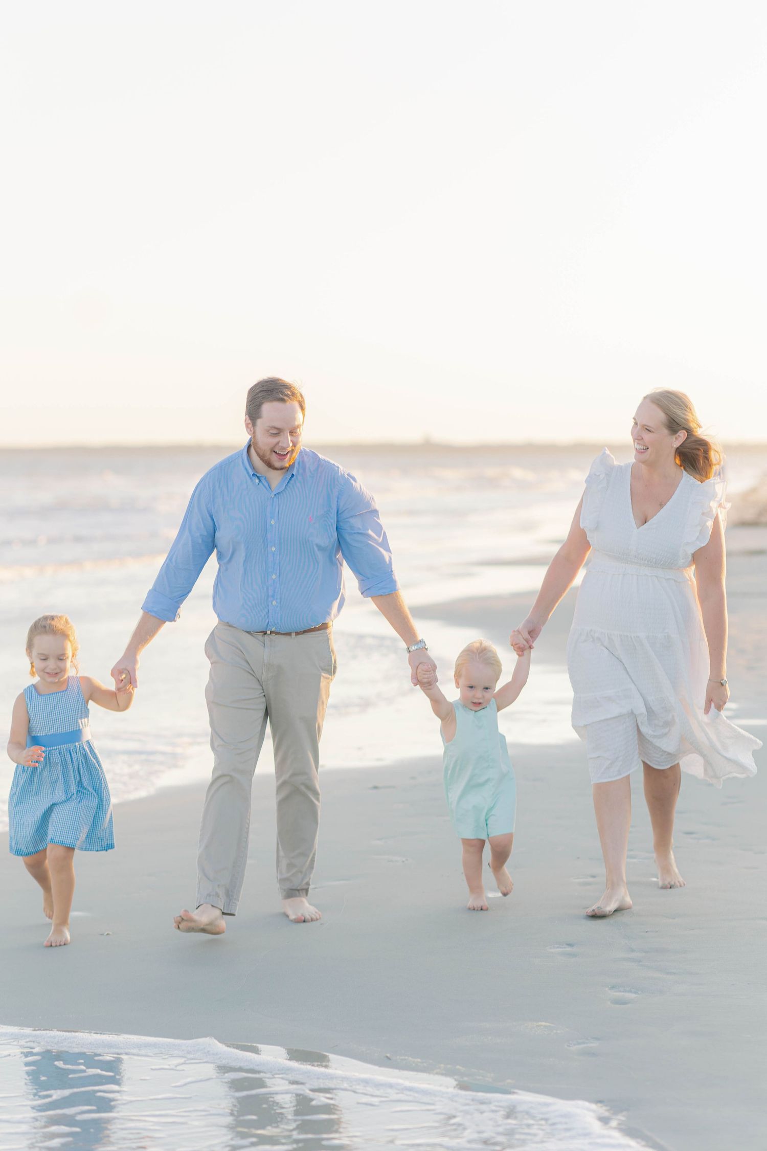 Family of four walking hand-in-hand on a sunny beach, parents and two young children dressed in blue and white.
