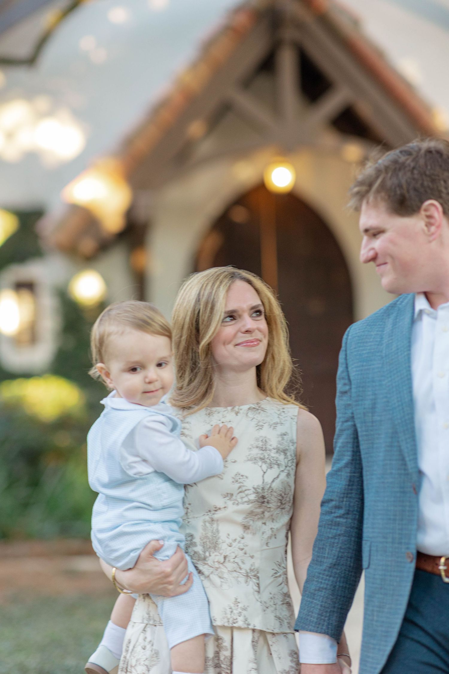 A smiling family with a woman holding a baby boy, standing beside a man in a blue suit near a charming lit venue.