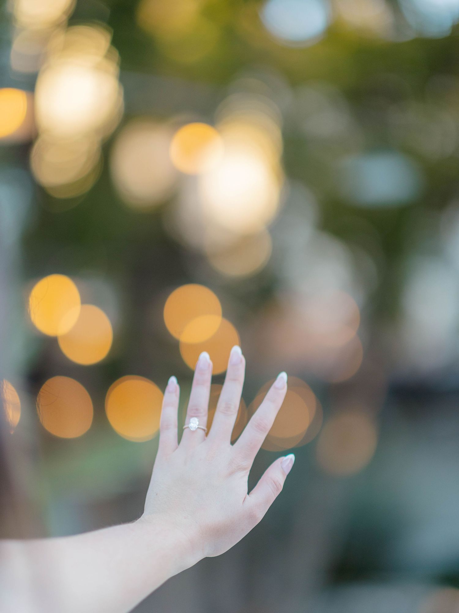 Close-up of a woman's hand displaying a diamond engagement ring with bokeh lights in the background.