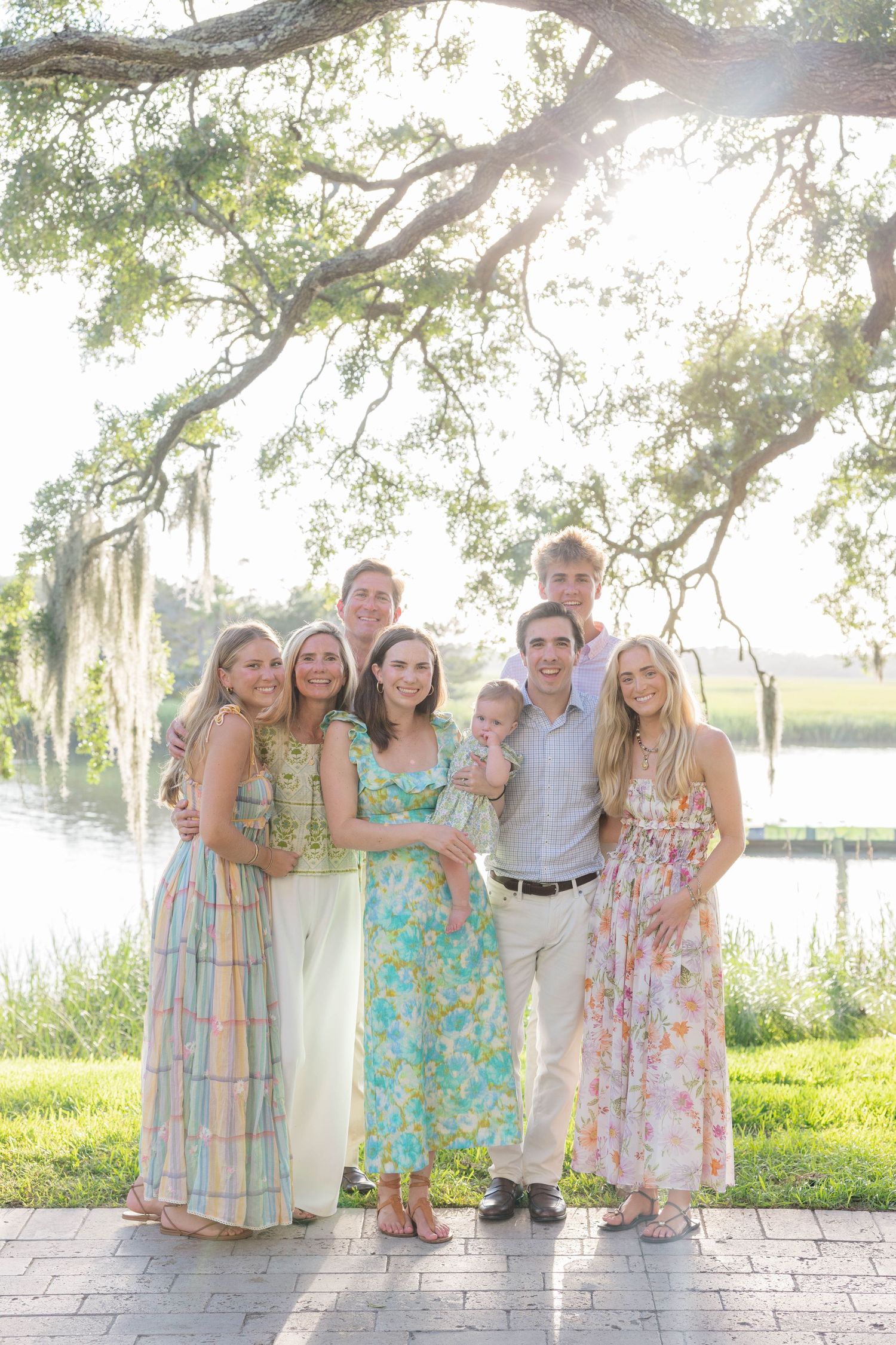 Large family group portrait outdoors near a lake, wearing pastel spring dresses and suits under a moss-draped oak tree.