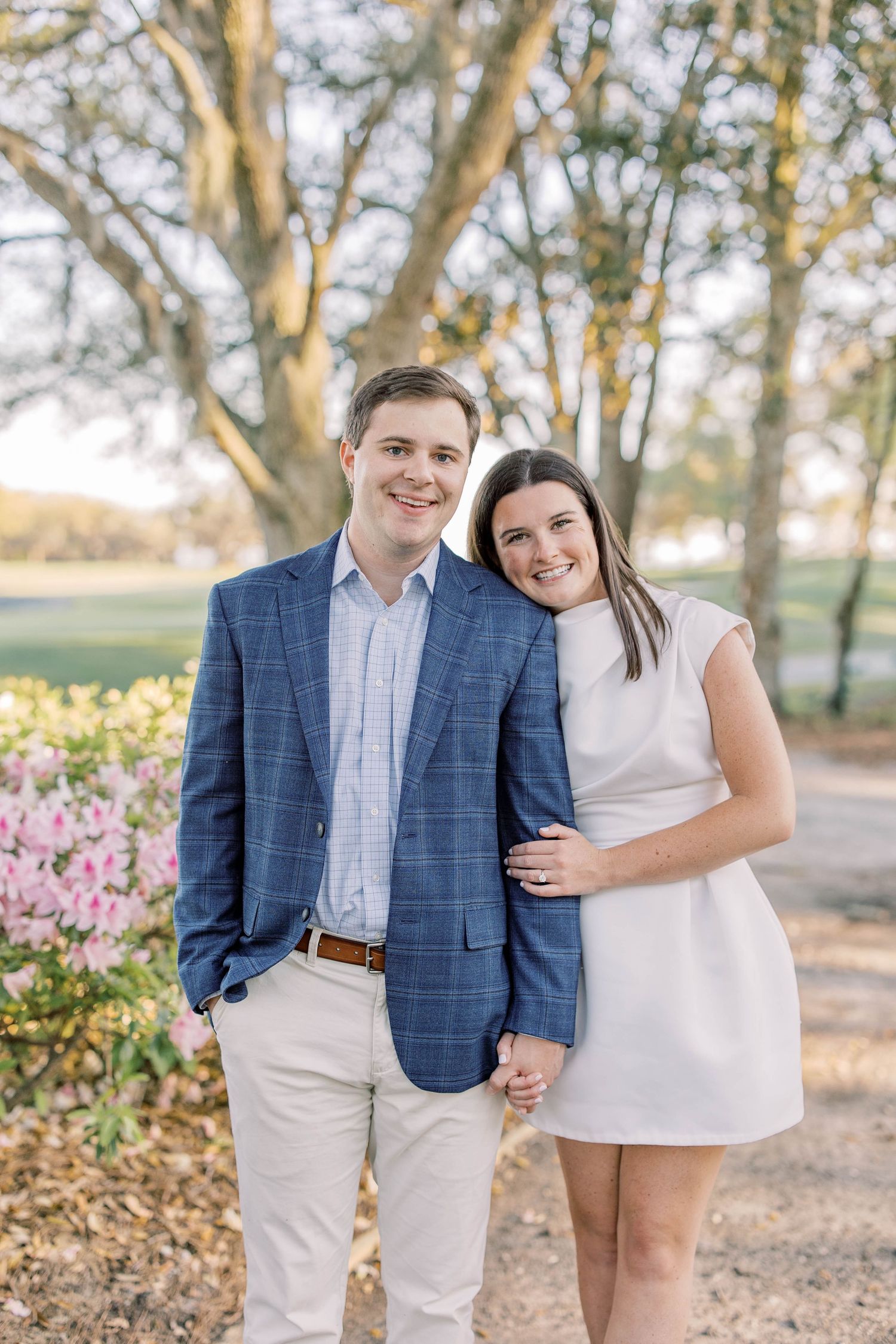 Smiling couple posing outdoors near pink flowers and trees, man in blue blazer, woman in white dress.