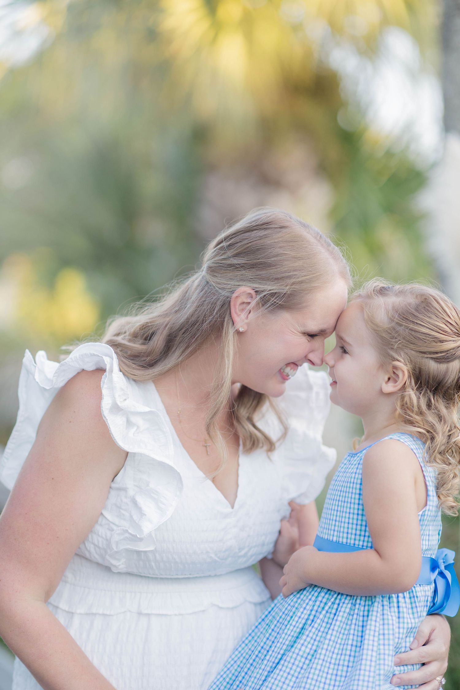 A smiling pregnant woman in a white dress touches foreheads with her young daughter in a blue gingham dress outdoors.