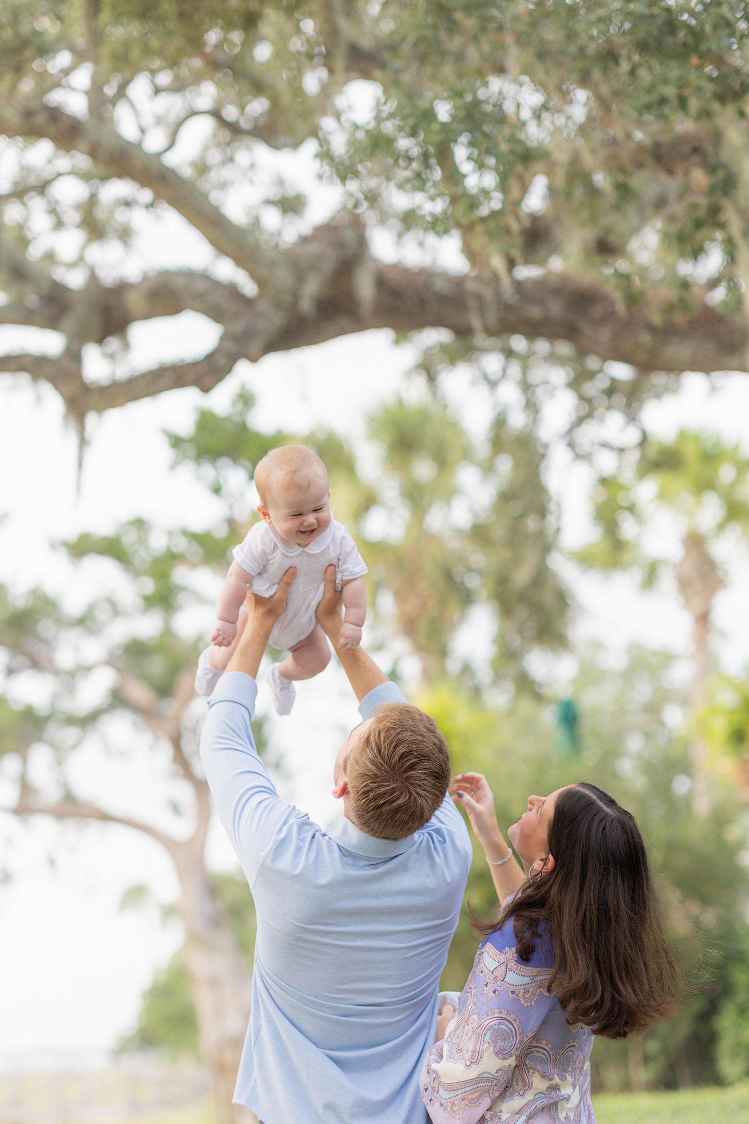 A joyful father lifts a smiling baby in the air outdoors while mother watches, surrounded by lush green trees.
