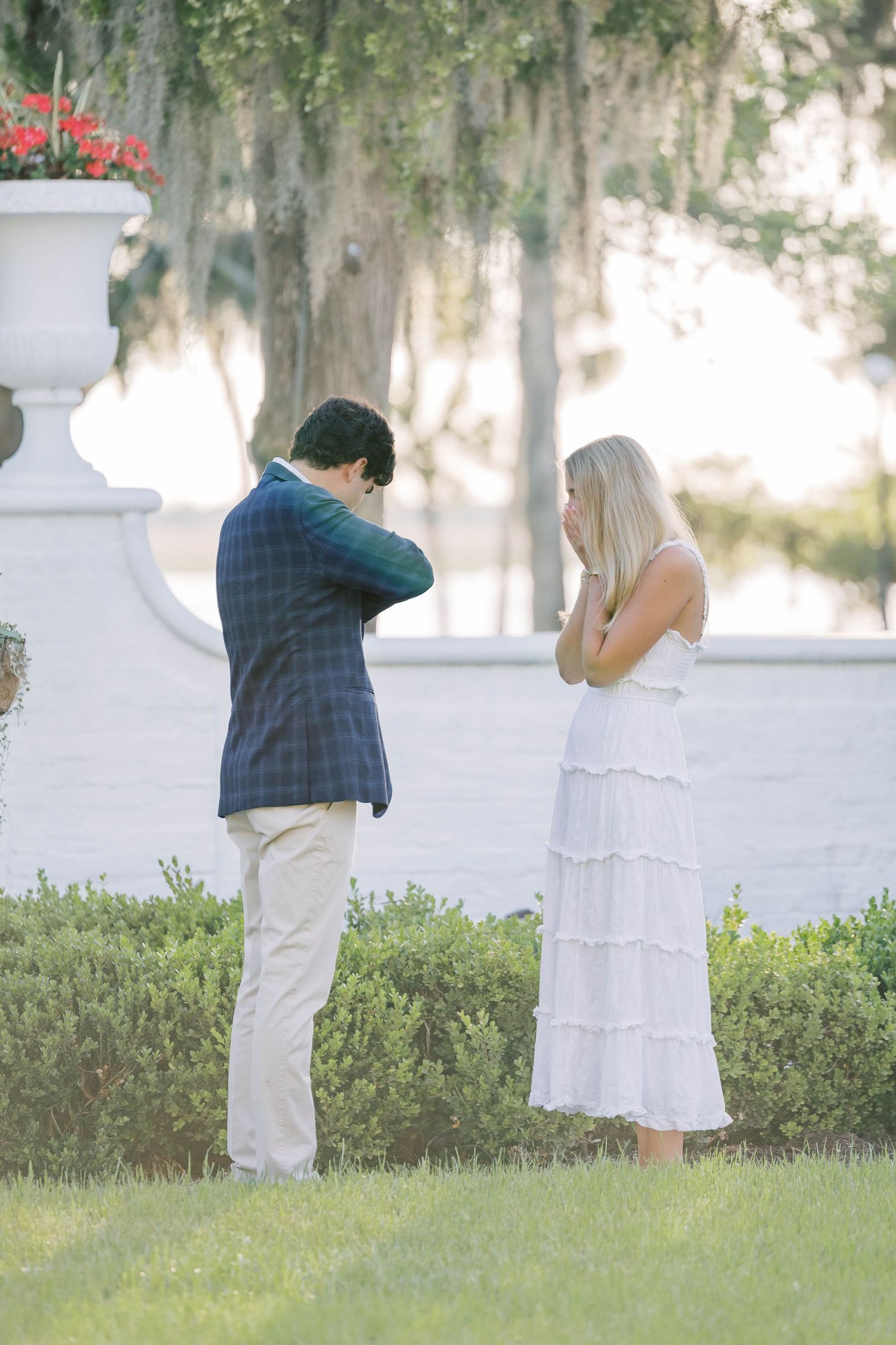 Man in plaid blazer and woman in white dress share an emotional moment outdoors near manicured hedges.