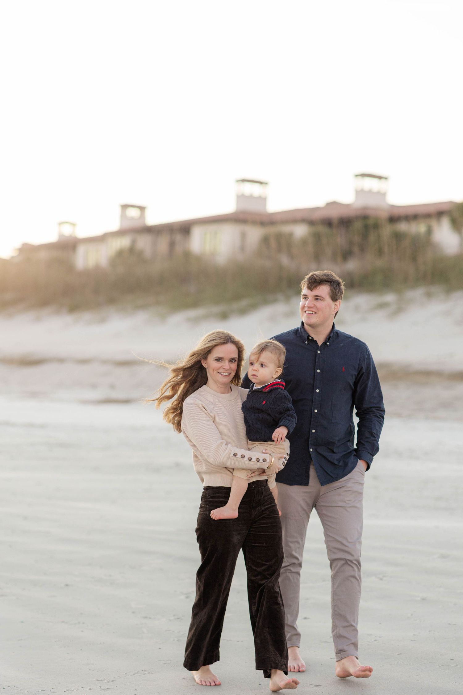 Young family with toddler posing on a sandy beach at sunset, woman holding child beside smiling man in navy shirt.