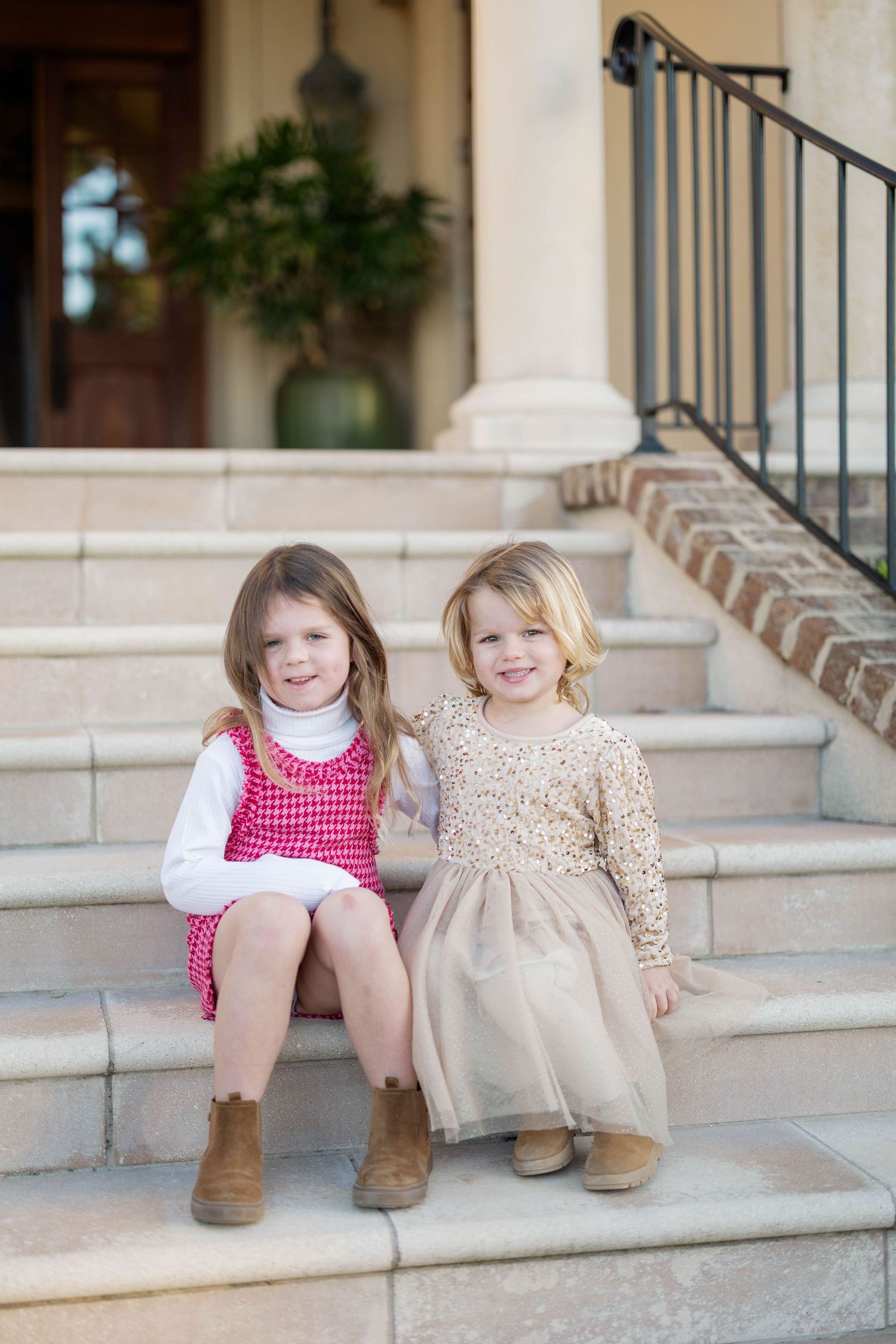 Two young girls in festive dresses sitting on stone steps, smiling, wearing brown boots outside an elegant home entrance.