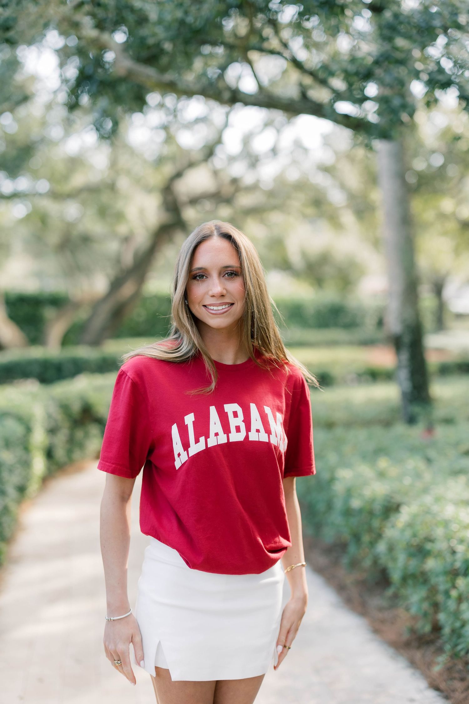 Woman smiling in red Alabama t-shirt and white mini skirt, standing on a garden path surrounded by green hedges.