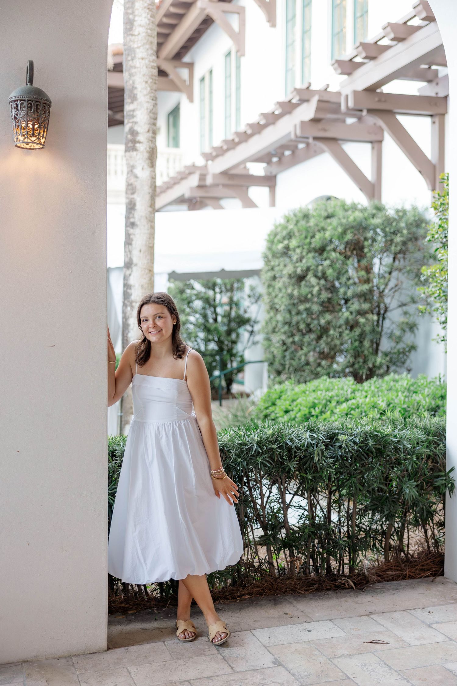 Young woman in white sundress smiling outdoors near white building with lush green hedges and decorative wall lamp.