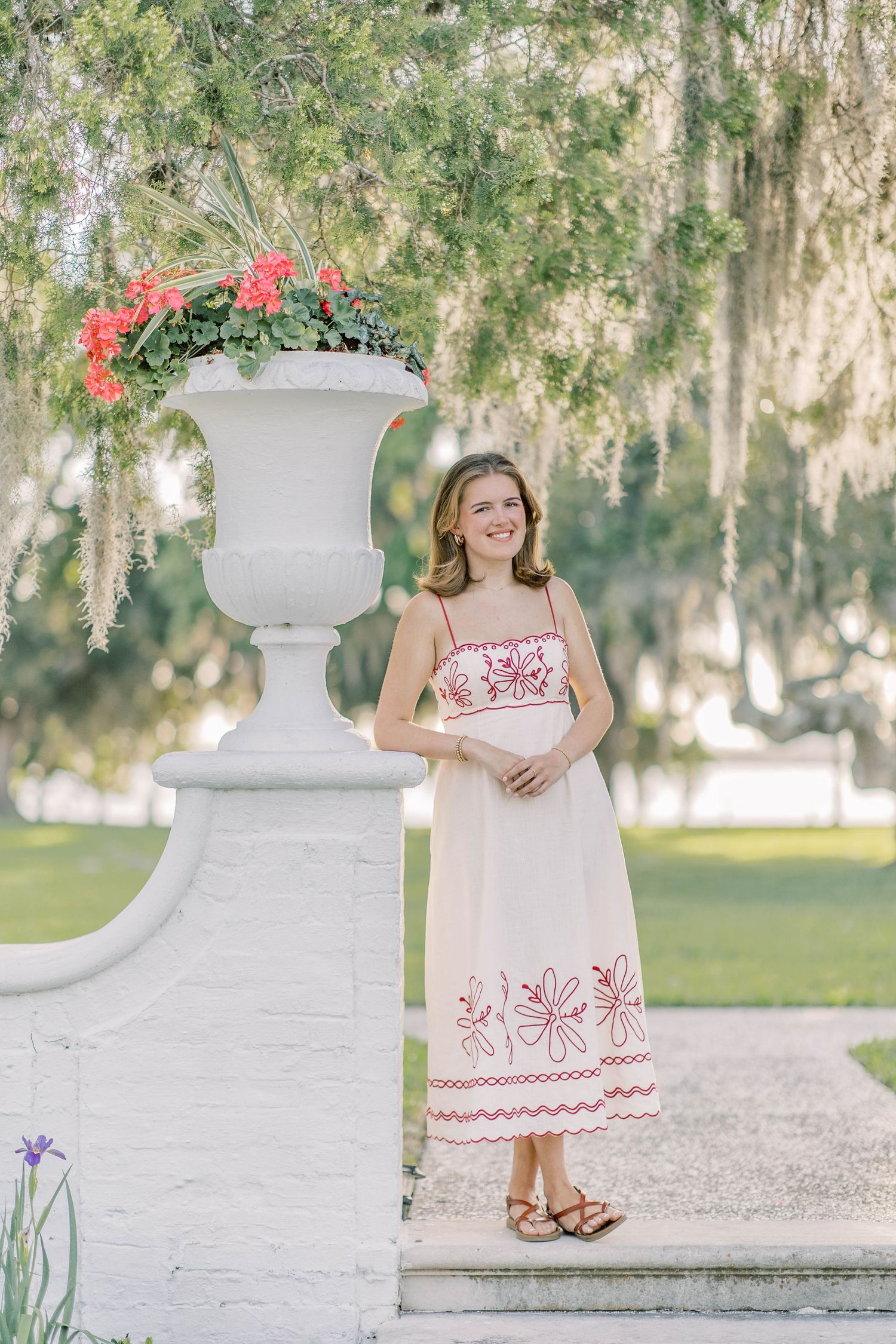 Young woman in white floral embroidered sundress smiling beside ornate garden urn with red flowers.
