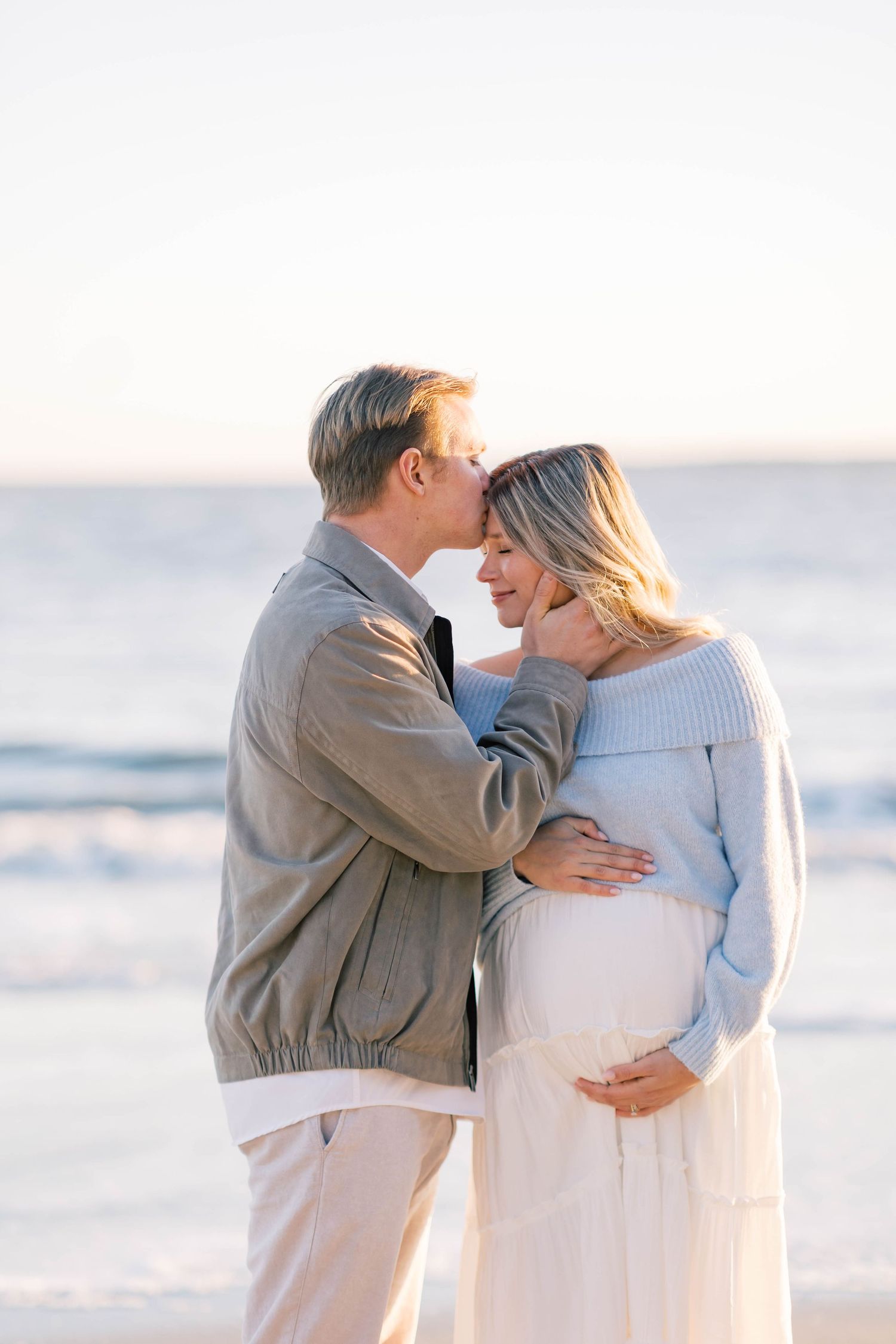 Couple sharing a tender moment on the beach, with the woman in a blue sweater cradling her baby bump at sunset.