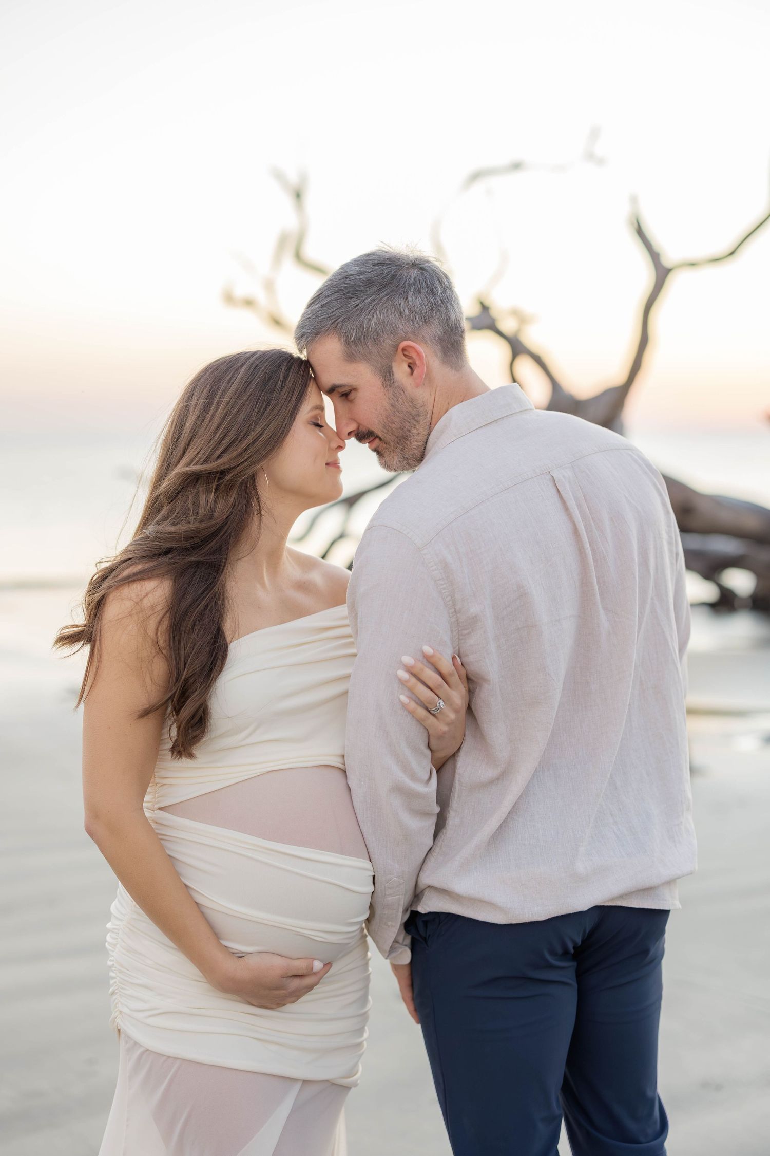 Pregnant couple sharing a tender moment on the beach at sunset, woman in white wrap dress, man in linen shirt.