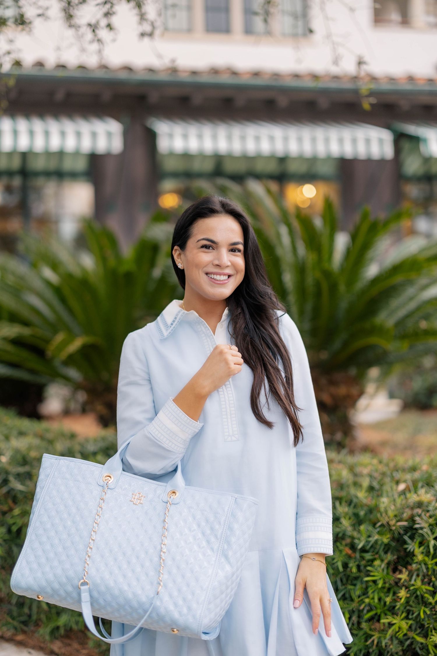 Smiling woman in light blue shirtdress carrying a quilted baby blue tote bag with gold chain straps outdoors.