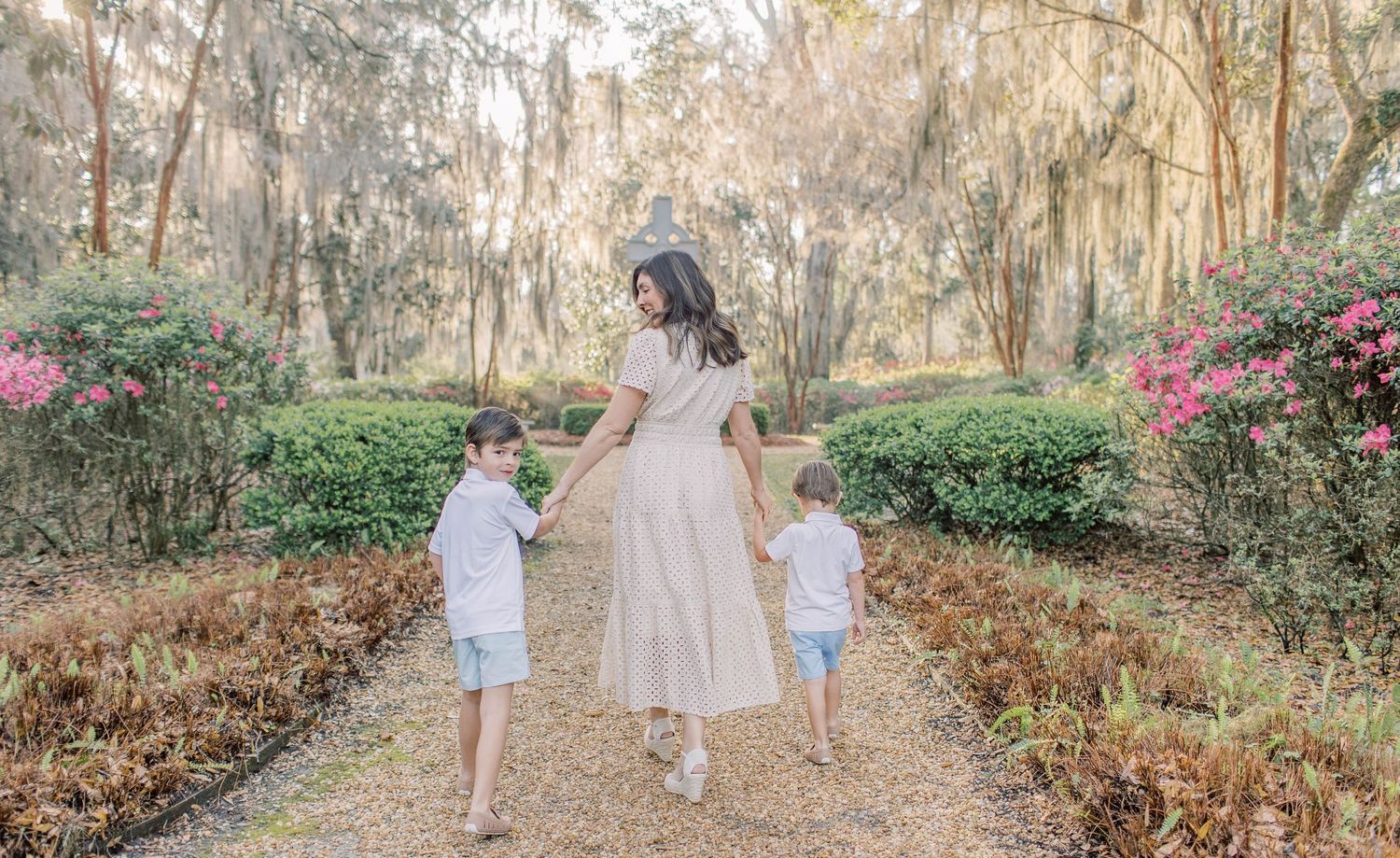 A mother in a flowy beige dress walks hand-in-hand with two young boys through a lush garden path.