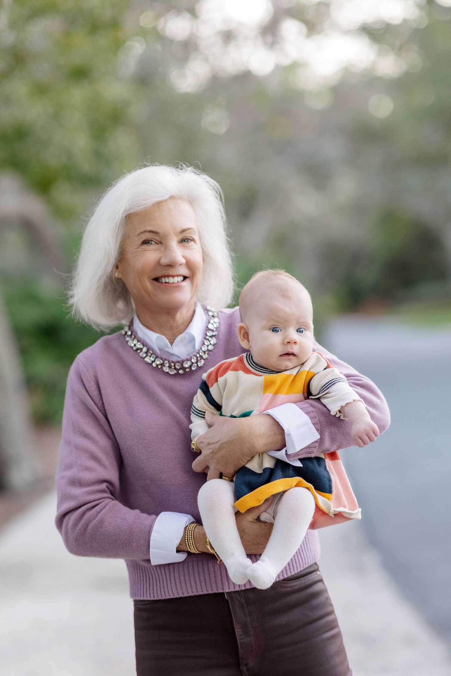 Smiling elderly woman with white hair holding a baby outdoors in a park setting.