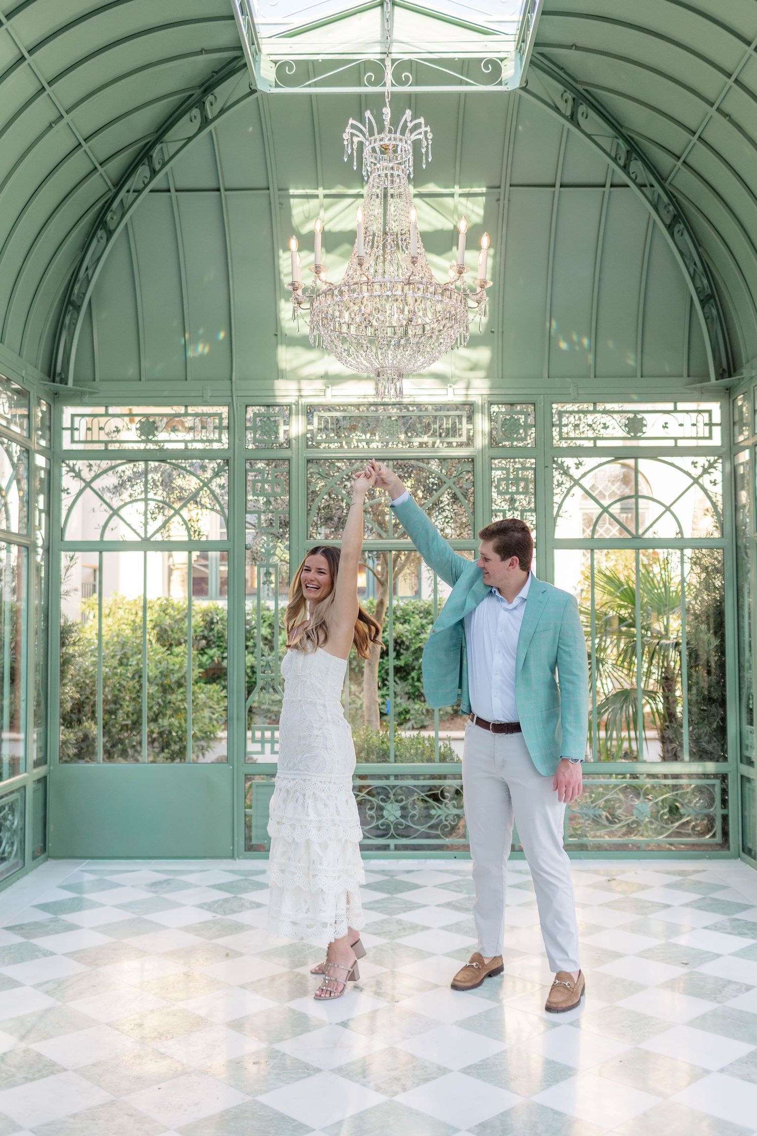 Couple dancing in elegant mint green glass conservatory with crystal chandelier and checkered marble floors.