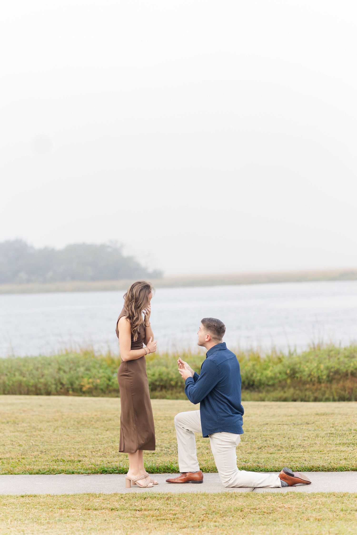 Man kneeling on one knee proposing to woman in brown dress by waterfront on grassy lawn.