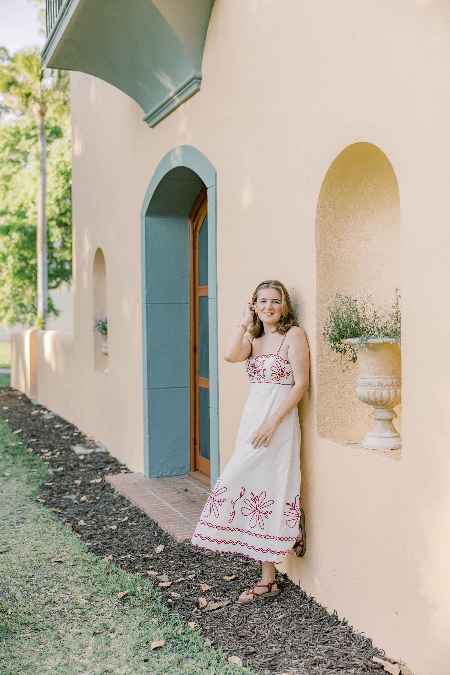 Young woman in white embroidered sundress posing by arched doorway of yellow Mediterranean-style building.