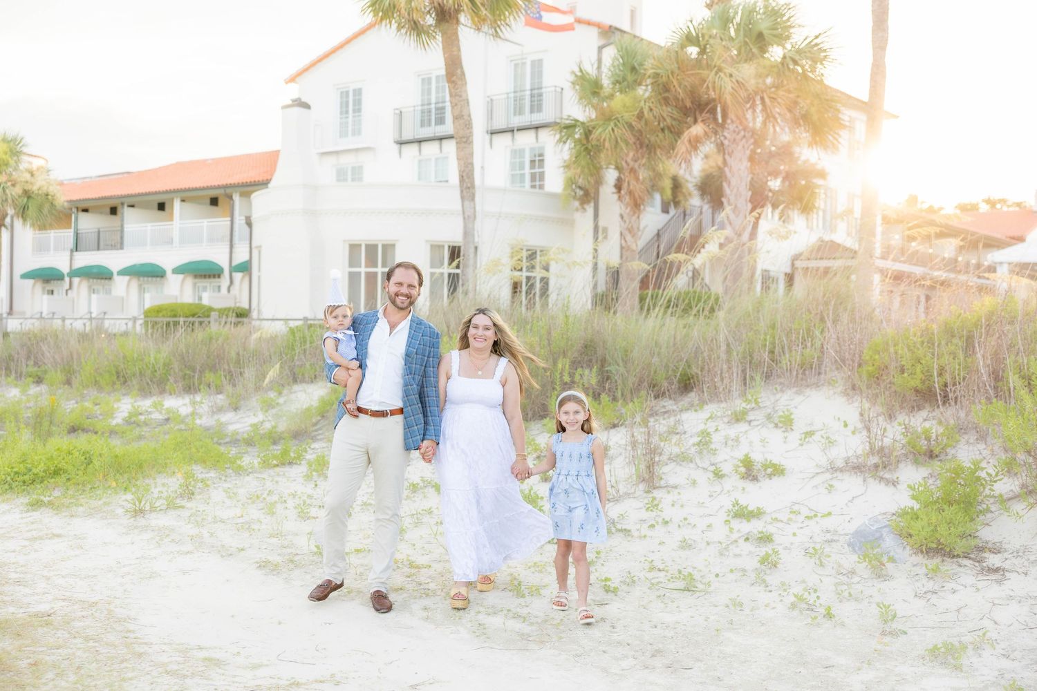 Family of three walking on white sand beach near a white coastal building with palm trees at golden hour.
