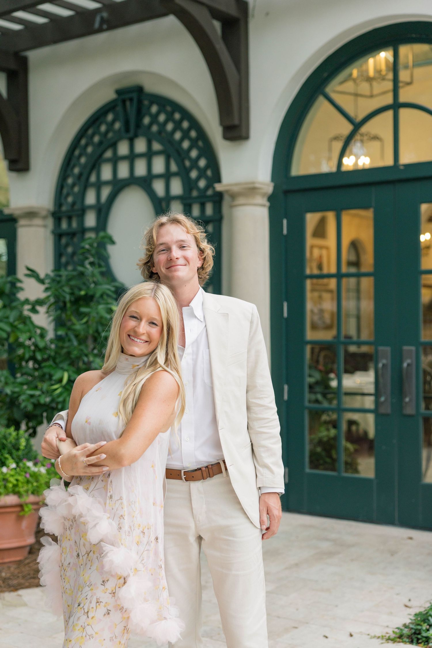 Smiling couple in elegant cream outfits posing before green arched doors at a upscale venue.