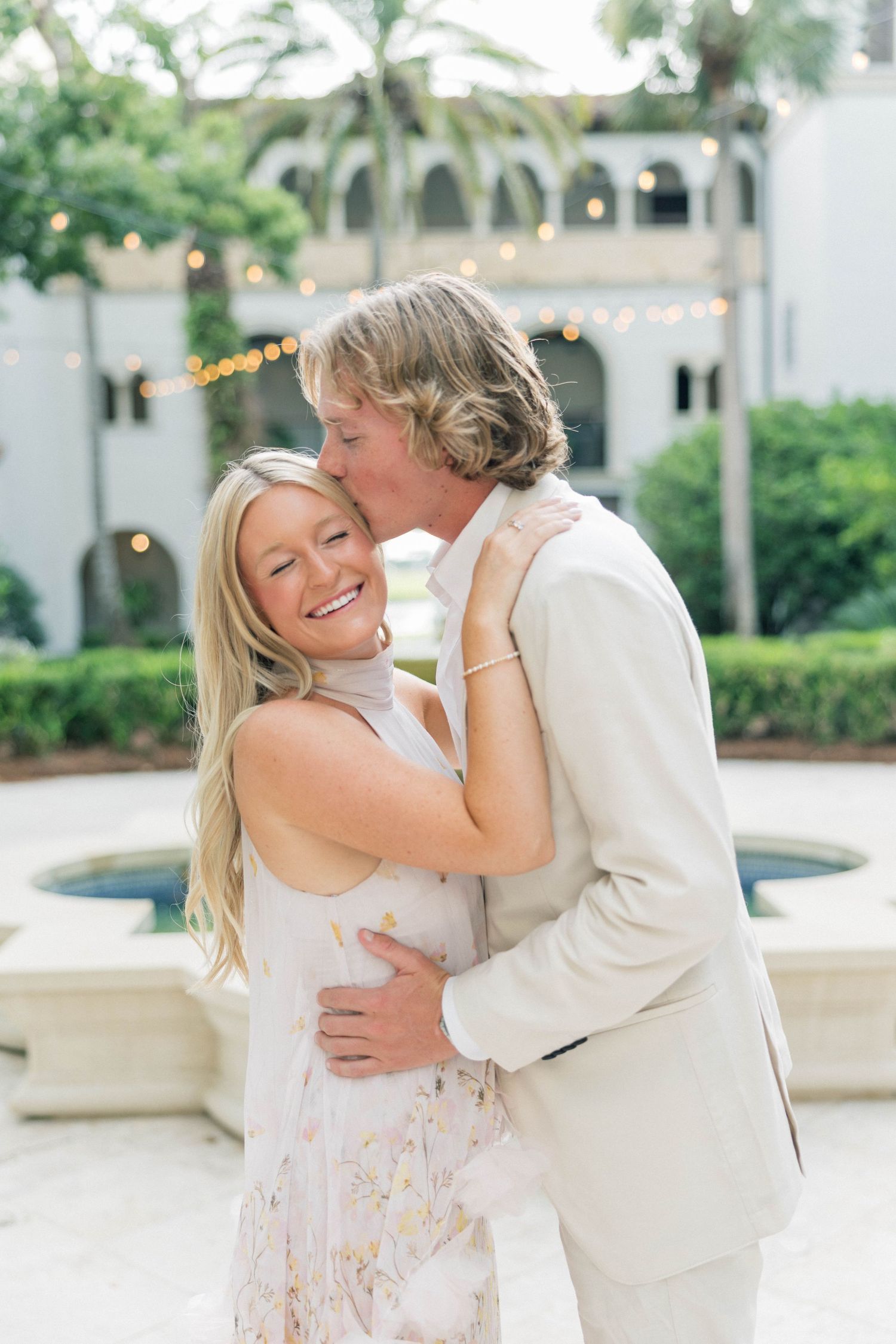 A smiling blonde woman in a floral dress receives a kiss from a man in a beige suit outdoors near a fountain.