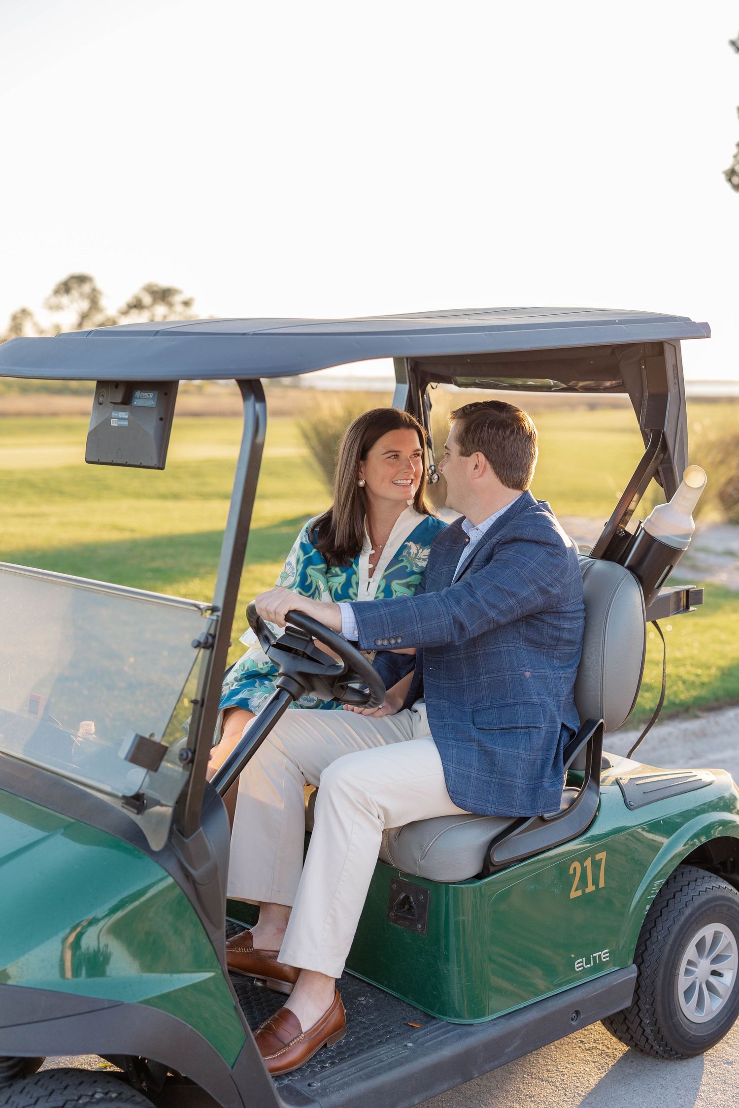 A couple sits together in a green golf cart numbered 217, smiling at each other on a sunny golf course.