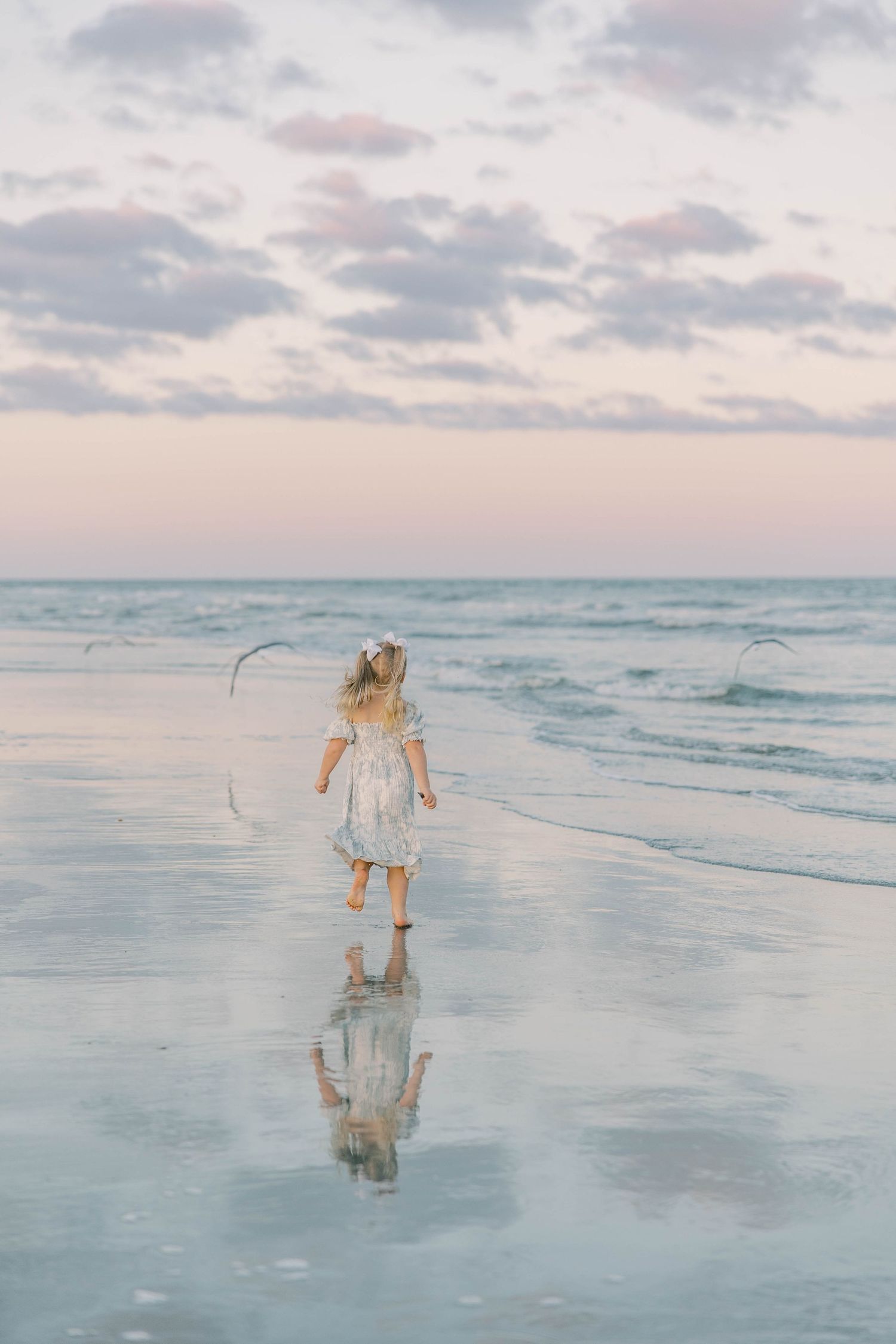 Young girl in floral dress walking barefoot toward ocean waves at a pastel sunset beach.