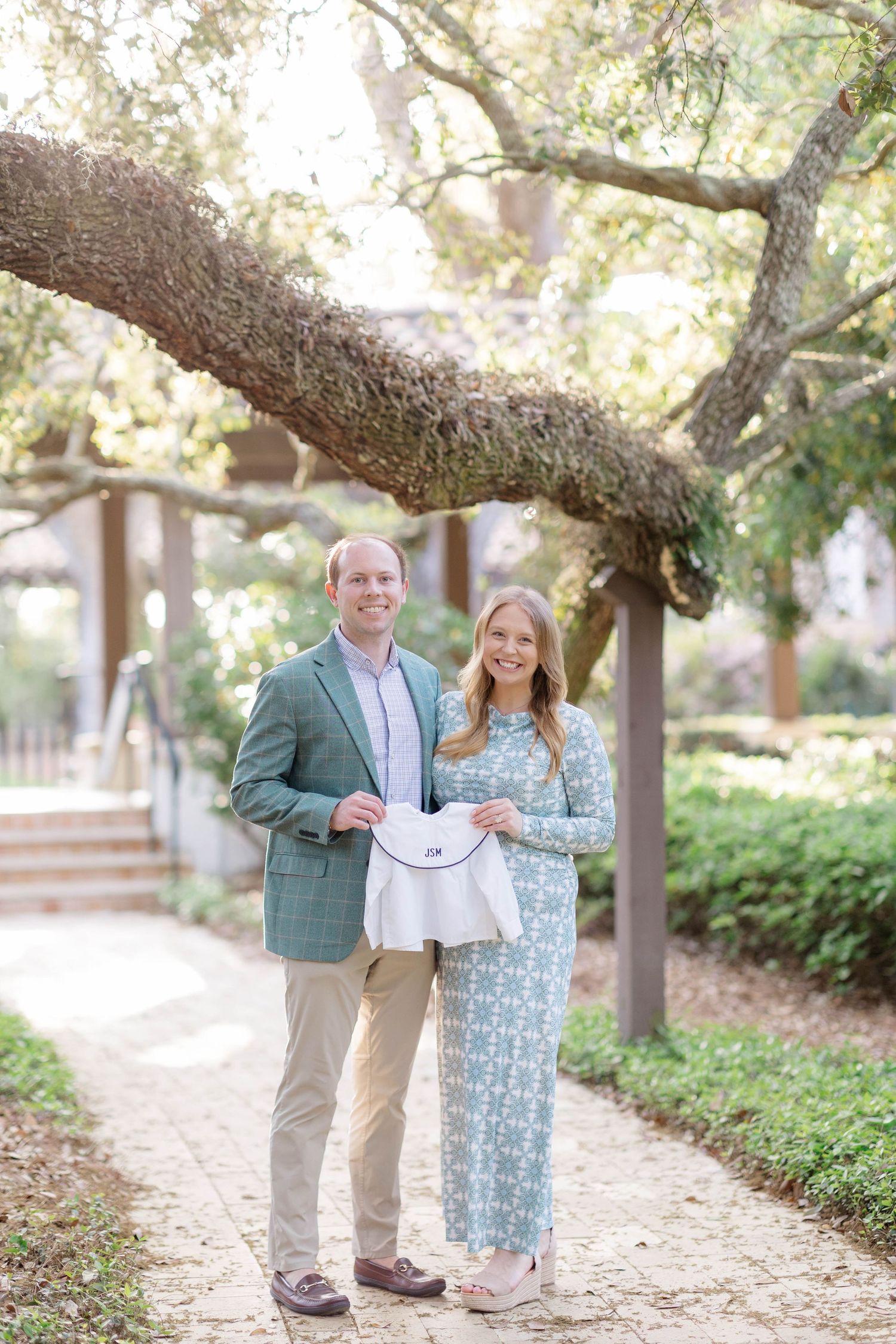 Couple smiling outdoors on a garden path under oak trees, holding a small white outfit for a baby announcement.