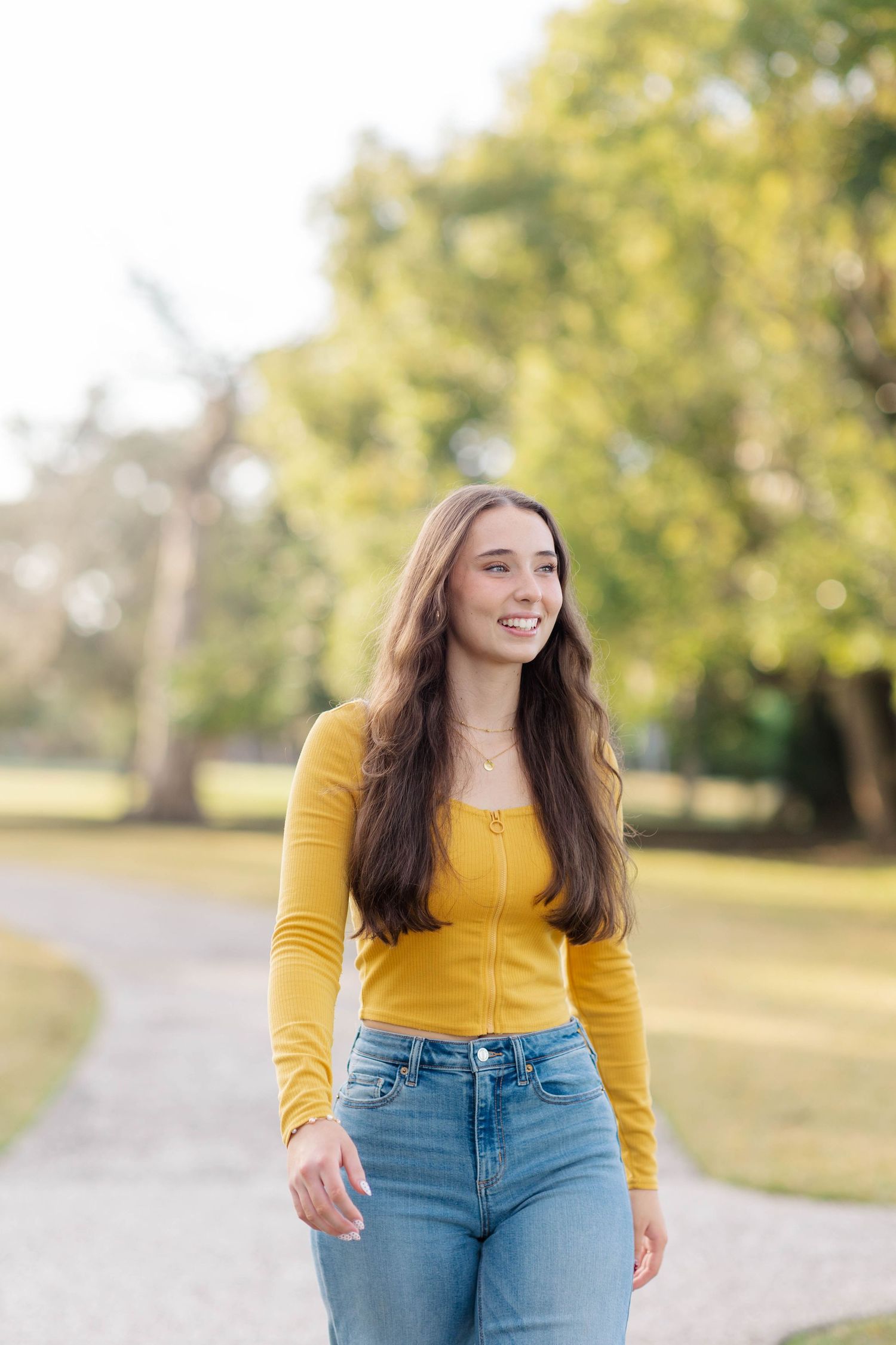 Young woman in yellow top and jeans smiling while walking on a park path with green trees in background.